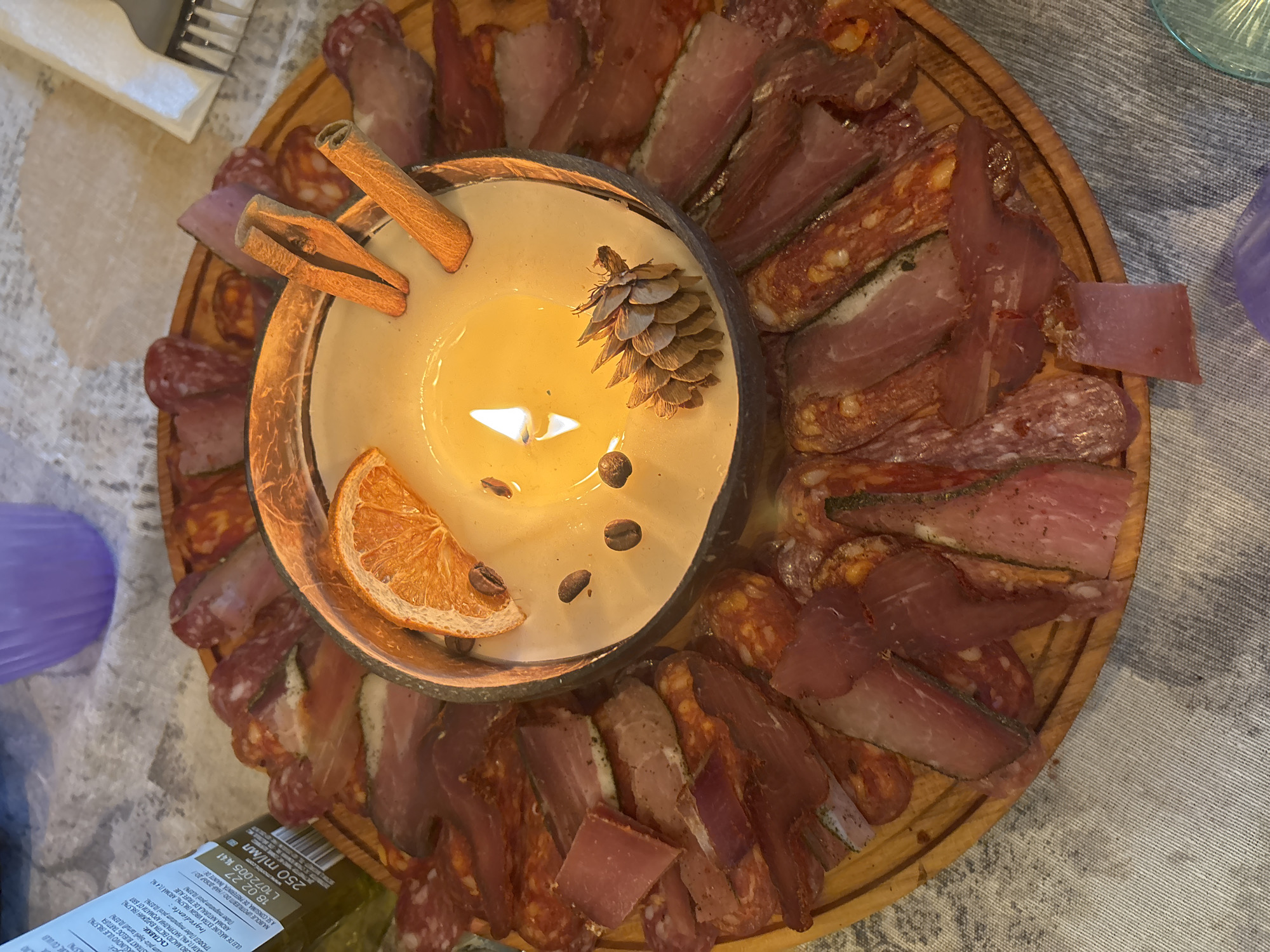 An overhead shot captures a decorative food and candle arrangement on a light-colored, patterned tablecloth, likely in Plovdiv, Bulgaria. At the center of a round wooden serving board is a lit candle in a rustic, dark-rimmed bowl, possibly a coconut shell. The candle's yellow wax is adorned with two cinnamon sticks, a small pinecone, a dried orange slice, and a few coffee beans, suggesting an aromatic and cozy ambiance.
Arranged circularly around the central candle on the wooden board is an assortment of sliced cured meats and sausages. These appear to be various types, some thinly sliced and others thicker, displaying different textures, colors, and marbling, indicating a charcuterie or antipasto platter.
In the upper left corner, silver forks are partially visible on a white napkin. Towards the left side of the frame, a translucent purple cup and a green bottle are present. The bottle has a label with text in multiple languages, primarily Romanian and Bulgarian, mentioning "ASLINOVO ULJE" (Olive Oil), "BUELOG TARTUFA" (White Truffle), "ULEI DE MASLINE EXTRA VIRGIN" (Extra Virgin Olive Oil), and "AROMA NATURALA DE TRUFE ALBE" (Natural White Truffle Aroma), along with the scientific name "Tuber magnatum pica." A "Best Before" date of "18.02.27," volume "250 ml/мл," and "www.monini.com" are also legible. The scene depicts a warm, inviting setting, likely for a meal or gathering, with the lit candle suggesting evening or an intimate atmosphere.