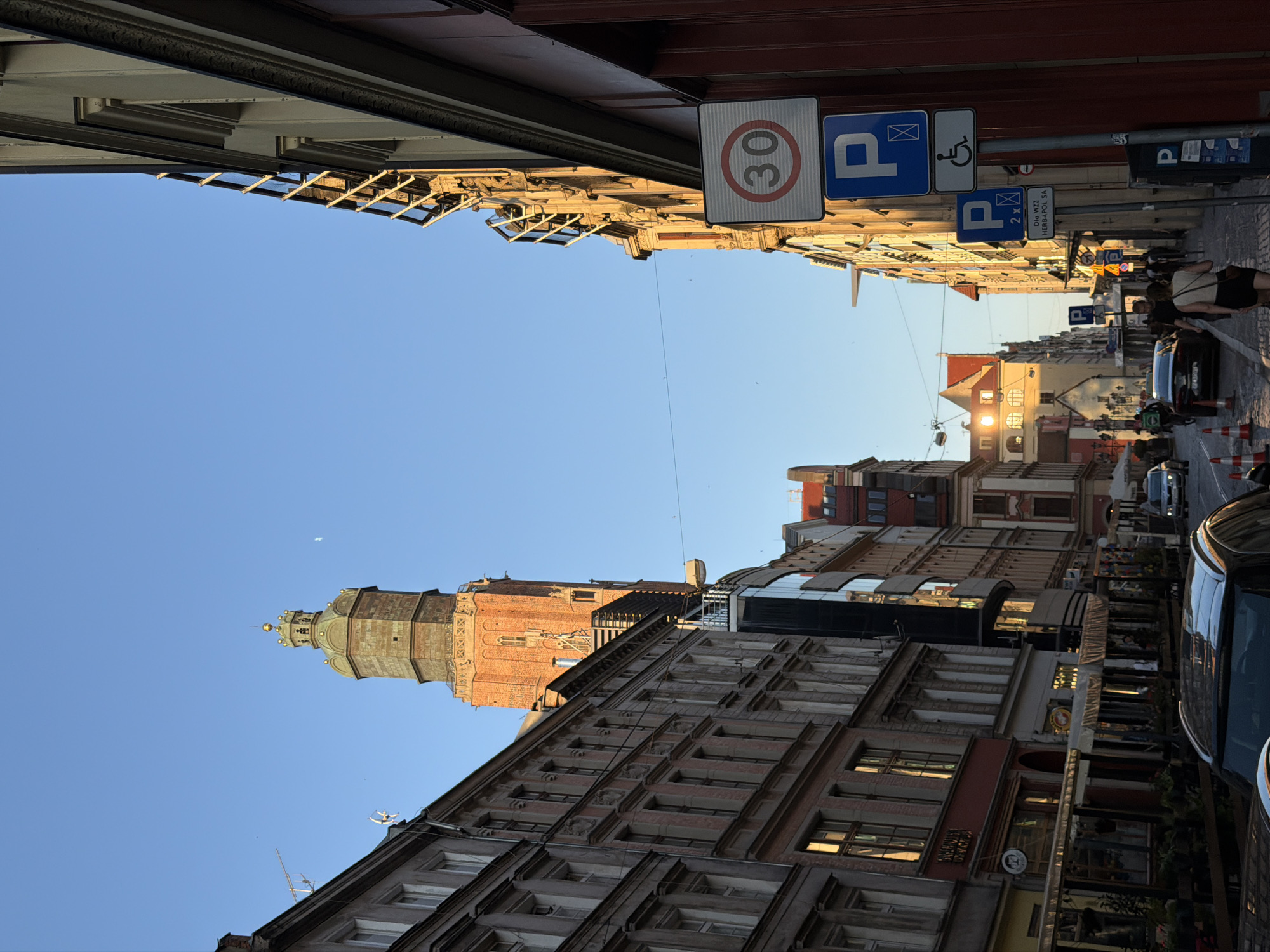 The image depicts a street scene in Wrocław, Poland, on a clear day with a bright blue sky. The foreground shows the roof of a black car. The street is lined with multi-story buildings, characteristic of European city architecture, with the left side featuring older buildings with detailed facades and numerous windows, some reflecting the sunlight. The right side of the street has buildings with more contemporary architectural elements, including exposed metal structures on the windows and a red facade.
Prominently in the background, a tall, brick tower with a green, ornate crown-like top rises above the buildings. This is likely St. Elizabeth's Church tower, a recognizable landmark in Wrocław. Further down the street, other buildings with pitched roofs and warm-toned facades are visible, suggesting a historic urban area.
Several traffic signs are present on the right side of the street: a speed limit sign indicating 30, two parking signs (one with a wheelchair symbol), and another parking sign with the text "Dla WZZ HERBAPOL SA".
On the street, there are a few cars, including a police car, and a small number of pedestrians are walking. The street is paved with cobblestones. Outdoor seating areas, possibly for restaurants or cafes, are set up in front of some of the buildings on the left side. The overall atmosphere suggests a vibrant urban environment during daylight hours, likely late afternoon given the warm light illuminating the buildings.