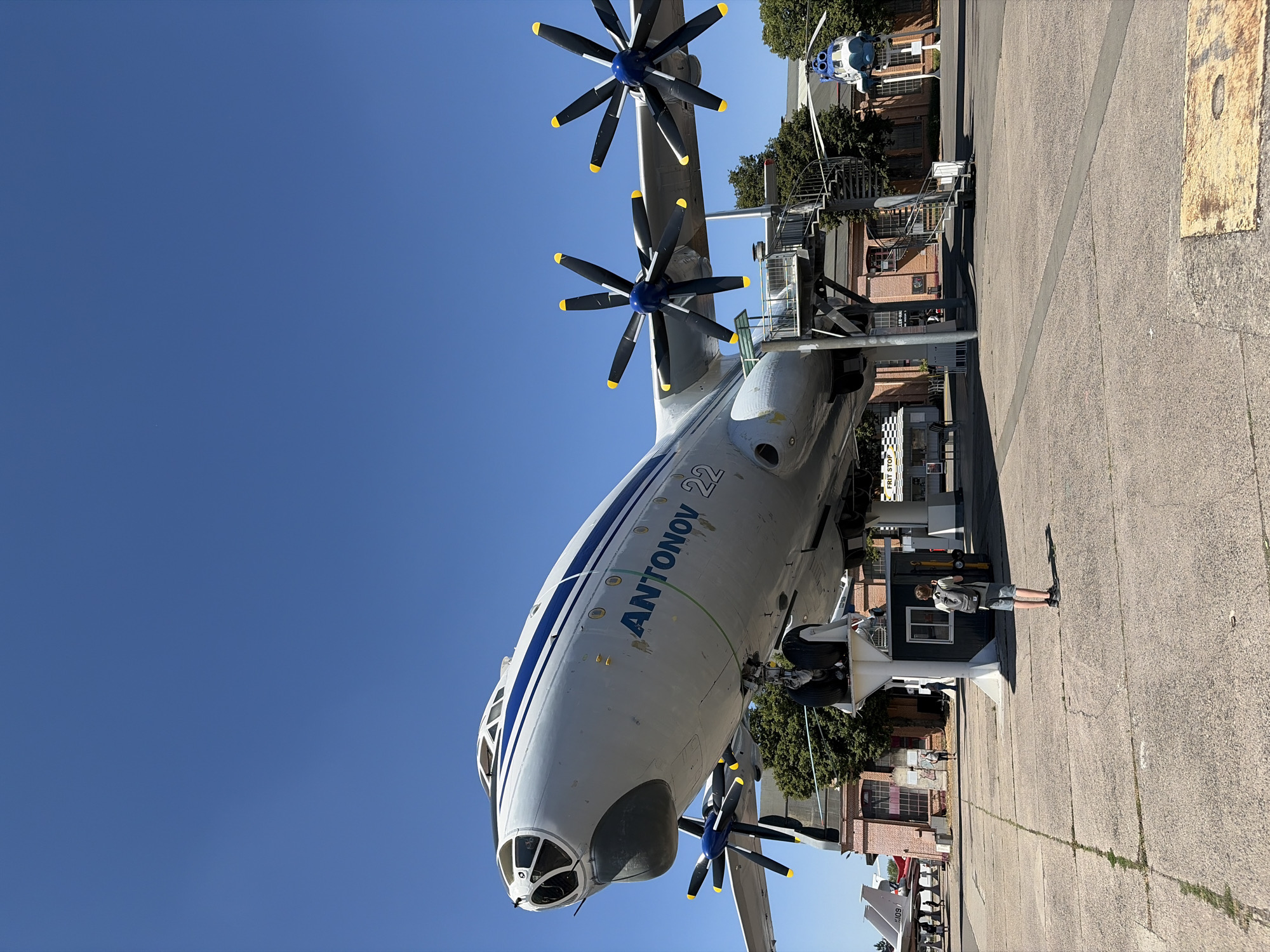 The image shows an Antonov An-22 aircraft displayed outdoors at the Technik Museum Speyer in Speyer, Germany. The large, four-engine turboprop aircraft is painted white with blue stripes along its fuselage and wings, and the text "ANTONOV 22" is visible on the side. The aircraft is elevated on large white supports, with a spiral staircase providing access to a walkway near its underside.
In the foreground, a young person wearing a grey backpack and shorts stands with their back to the camera, looking up at the aircraft. Other people can be seen in the background near other exhibits and buildings. To the right of the An-22, a blue and white helicopter is suspended in the air. The ground is paved with concrete, showing some cracks and weeds. Behind the aircraft are brick buildings, trees, and a clear blue sky. A small food stand labeled "FRIT STOP" is visible to the left of the aircraft's support structure. Several other aircraft, including what appears to be a Tupolev Tu-144, are parked in the distance.