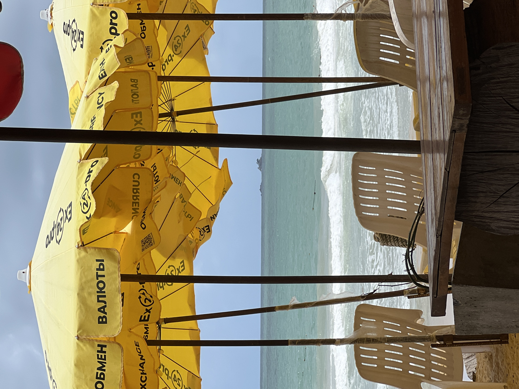 The image depicts a beach scene in an unknown location. The foreground features yellow beach umbrellas with exchange rate information in Russian ("ОБМЕН ВАЛЮТЫ" - "Currency Exchange"). Behind the umbrellas, there is a view of the ocean with visible waves and a light blue sky. A wooden table and stacked white plastic chairs are partially visible in the right foreground, suggesting a beachside cafe or restaurant.