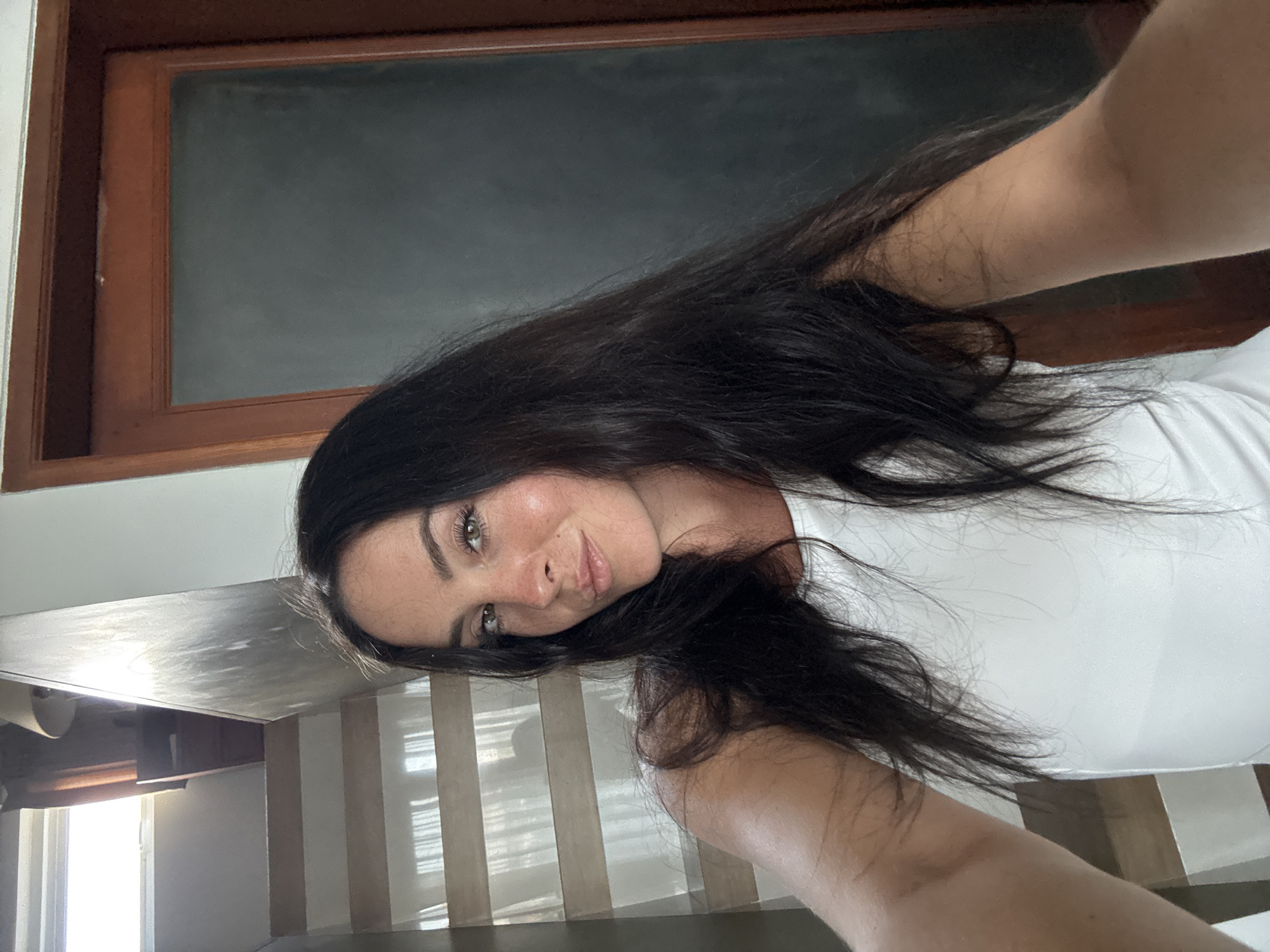In a selfie taken indoors, a woman with long, dark, wavy hair is positioned in the center of the frame, facing the camera. She has light skin, green eyes, and is wearing a white tank top. Her head is slightly tilted to the right. Behind her, a wooden door with a dark glass pane is visible on the right side of the image. To the left, a staircase with light-colored steps and a white railing leads upwards. The background also shows a window with blinds and a lamp on a shelf. The lighting suggests it is daytime, with natural light illuminating the scene. This image was taken in North Kuta, Indonesia.