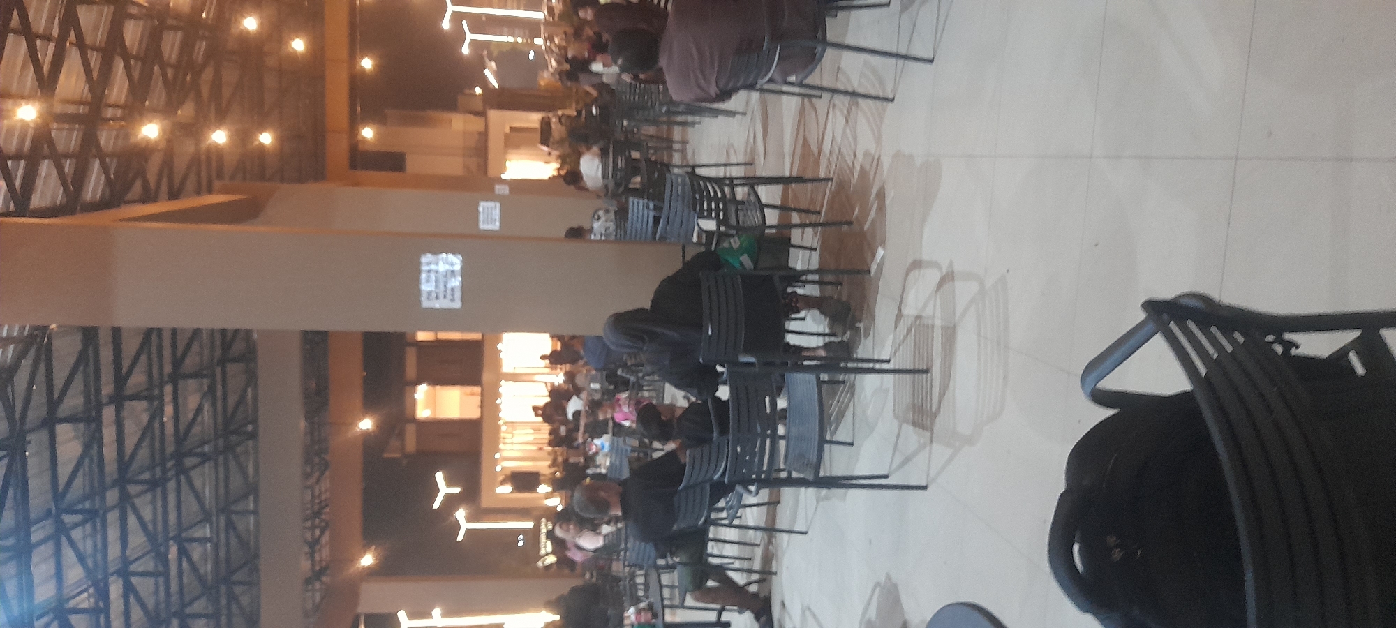 The image depicts an indoor gathering space, likely a cafe or restaurant, with a high ceiling featuring exposed metal beams and multiple light fixtures. The environment is illuminated by warm artificial lighting. In the foreground, several black metal chairs are arranged around tables, some of which are occupied by people. The floor is tiled in a light beige color.
In the mid-ground, a large cylindrical pillar is visible, painted in a light brown hue. On this pillar, there are two white signs with text and graphics that are not clearly discernible due to image resolution. Beyond the pillar, more tables and chairs are arranged, with a diverse group of people seated and socializing. Some individuals are wearing headscarves. In the background, a brightly lit building facade with large windows suggests an outdoor or open-air section of the establishment. The overall atmosphere appears to be casual and communal, with a moderate number of patrons present. The specific location within Kragilan, Indonesia, is not explicitly indicated by any visual cues in the image.