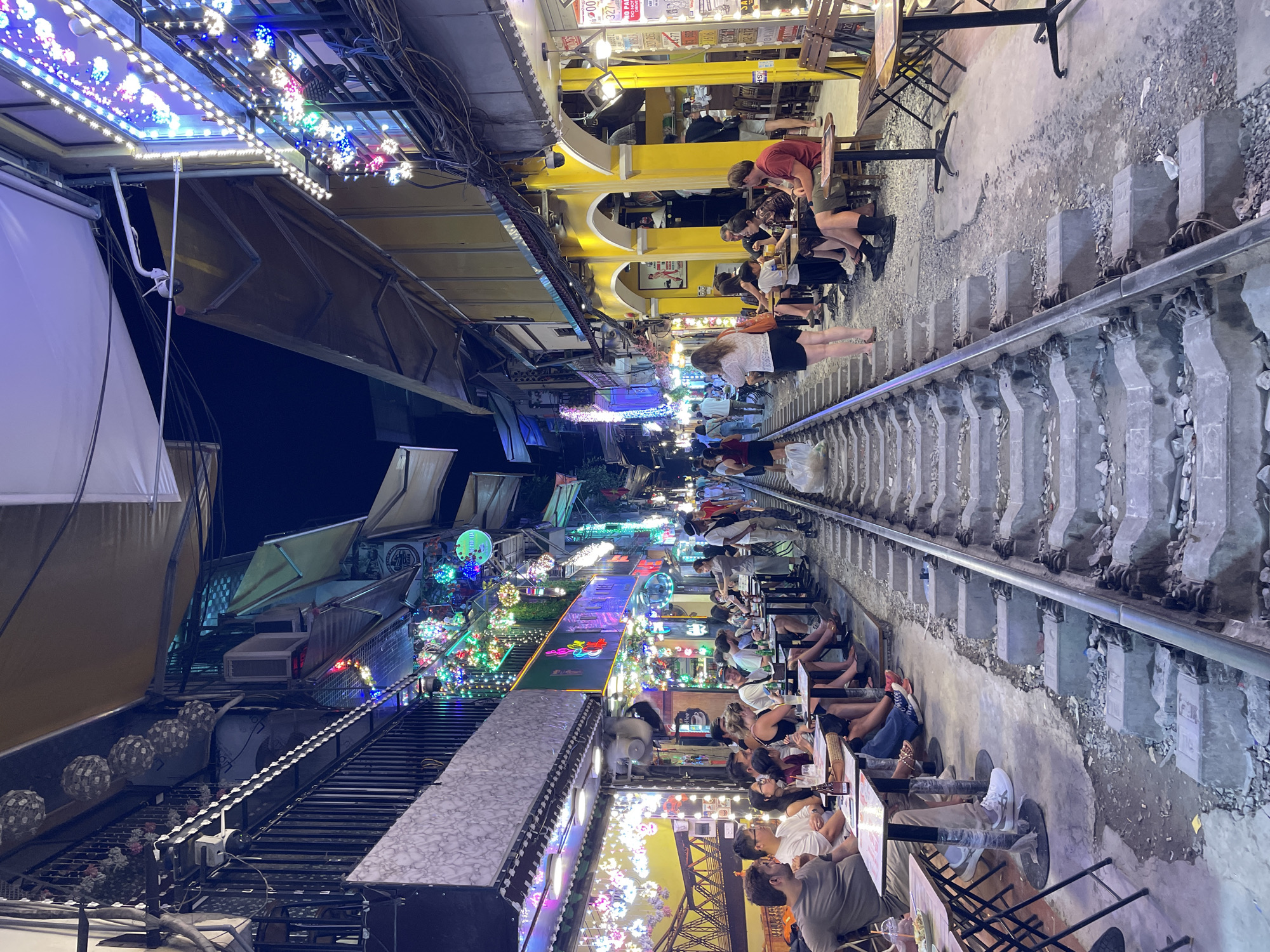 The image depicts a vibrant street scene in Hà Nội, Vietnam, specifically along the Hà Nội train street. Numerous people are seated at tables set up on either side of active railway tracks, suggesting a unique dining or drinking experience. The street is illuminated by strings of colorful decorative lights draped across buildings and awnings, creating a lively and festive atmosphere. Several buildings lining the street feature businesses with open fronts, and some display signage. The railway tracks themselves are a prominent feature, running through the middle of the scene and serving as a thoroughfare for pedestrians and the dining establishments. The time of day appears to be evening or night, given the artificial lighting. The overall impression is one of a bustling, urban environment adapted for tourism and commerce.