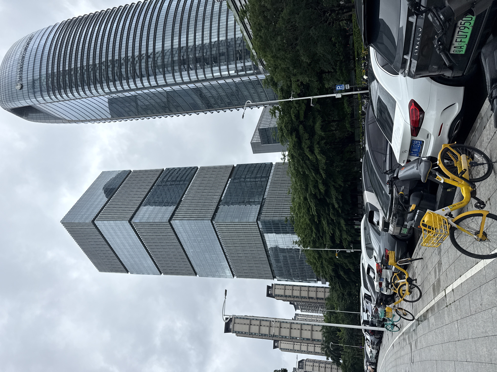 This is a vertical, eye-level, outdoor shot of an urban scene on a cloudy day. The image features modern architecture, including a curved glass skyscraper in the upper left, and a rectangular building with a stacked, geometric facade in the center. In the foreground, a row of parked cars and yellow and turquoise shared bicycles lines a sidewalk. The cars are modern, including a white Mercedes-Benz. The bicycles are equipped with baskets and appear to be part of a bike-sharing service. Lush green trees are visible behind the parked vehicles and bicycles, separating the street from other buildings in the background. These background buildings are tall and appear to be residential complexes. A parking sign is visible on a pole. Text on a license plate is visible on the white Mercedes-Benz, reading "T N102B". Another license plate on a black vehicle shows "粤AFU7950". The overall impression is of a modern, organized city environment with accessible transportation options.