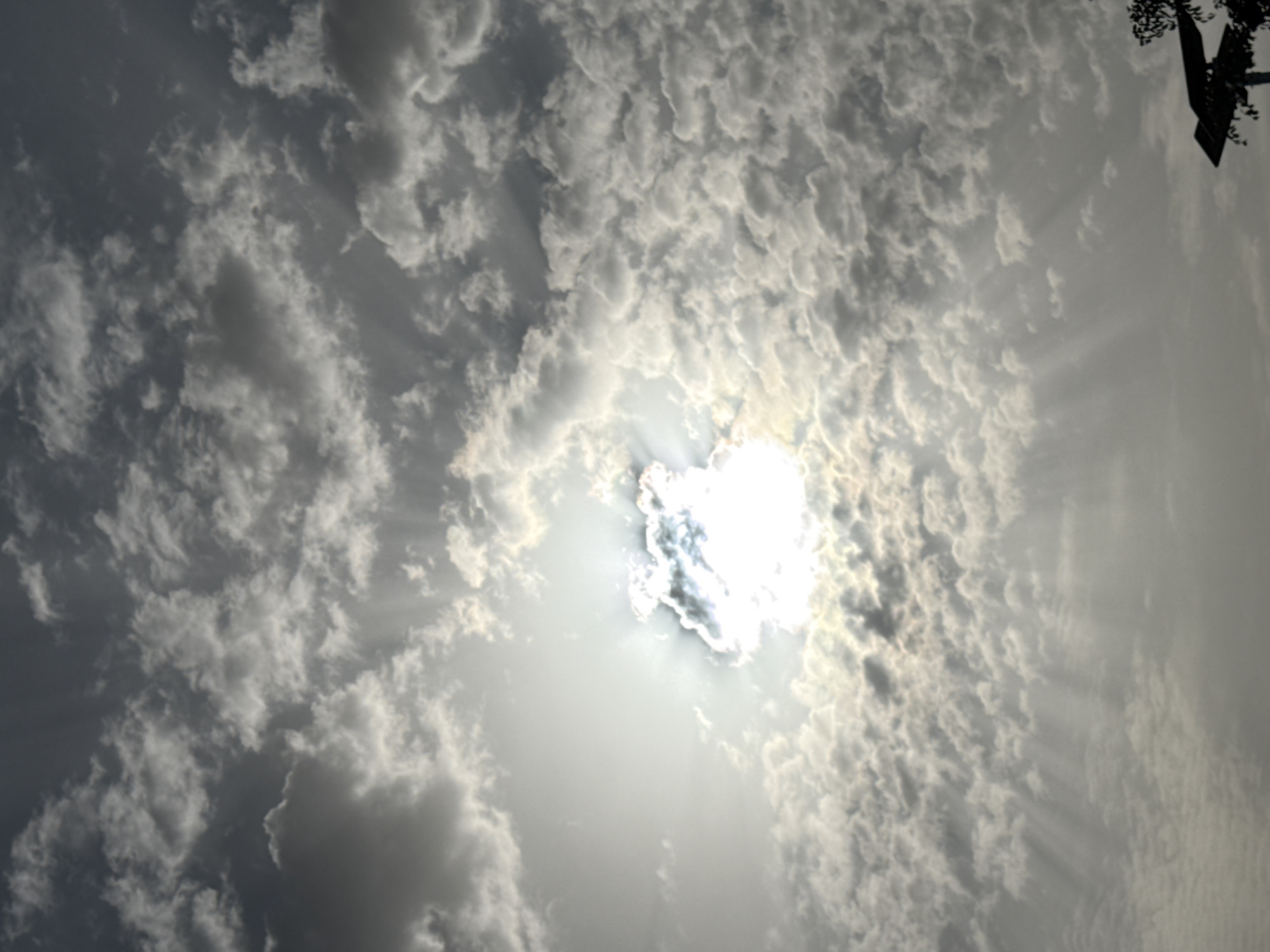 The image shows a sky filled with textured clouds, with the sun shining brightly through a break in the clouds. The light rays from the sun are visible, piercing through the cloud cover. In the top right corner, there is a dark object, possibly a tree branch or a man-made structure. The scene appears to be outdoors, during daylight. Specific location in Ado, Nigeria cannot be determined from the image alone.