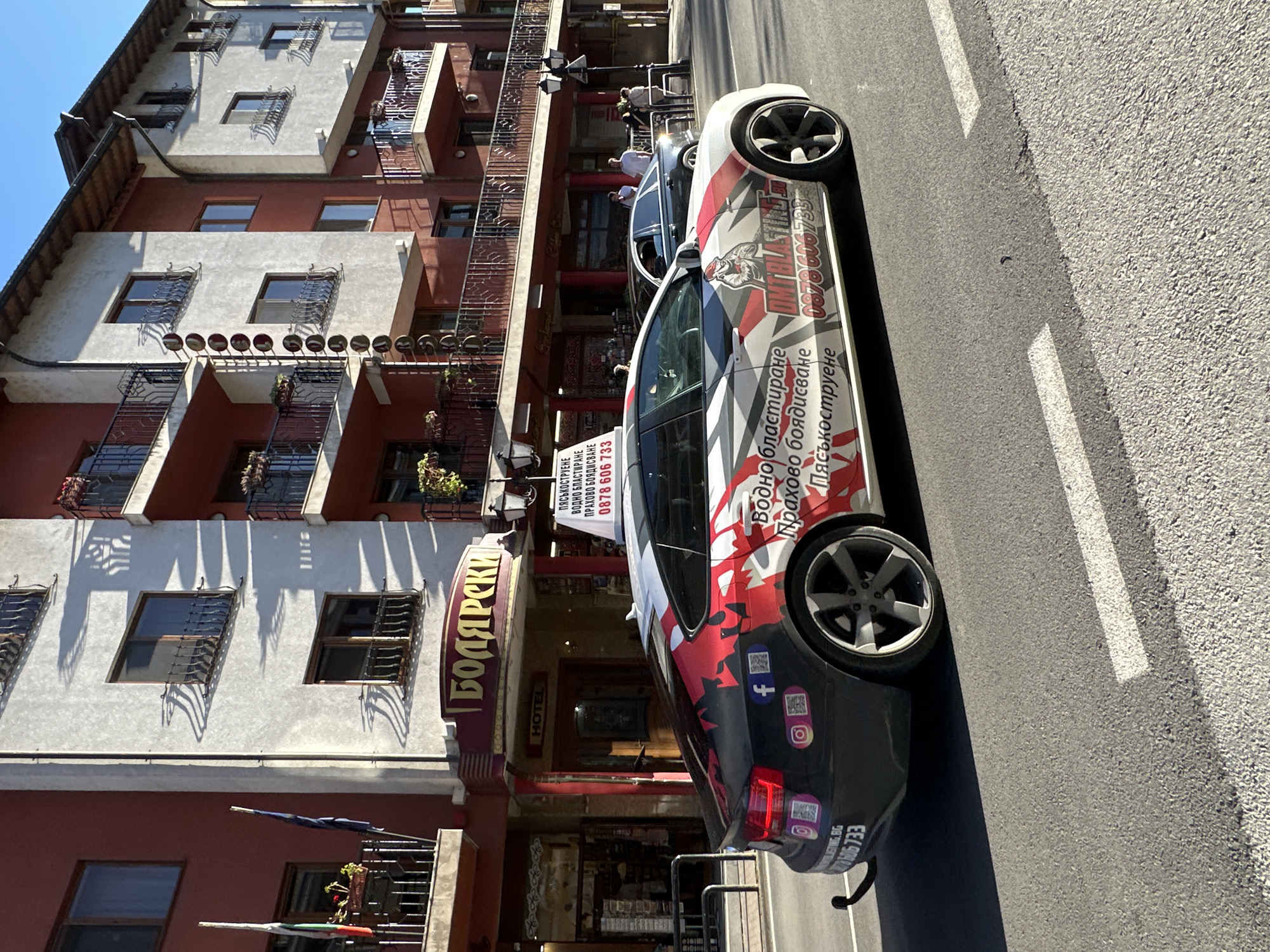 A black and white Audi A7 is parked on a street in Veliko Tarnovo, Bulgaria, in front of a building with a sign that reads "БОЛЯРСКИ" and "HOTEL". The car is decorated with red and white graphics and text, including a phone number. Several QR codes and social media icons are also visible on the car's rear. People are walking in the background, and a dark sedan is parked further down the street. The scene appears to be a sunny day in an urban environment, possibly a commercial district or a street lined with hotels and shops.