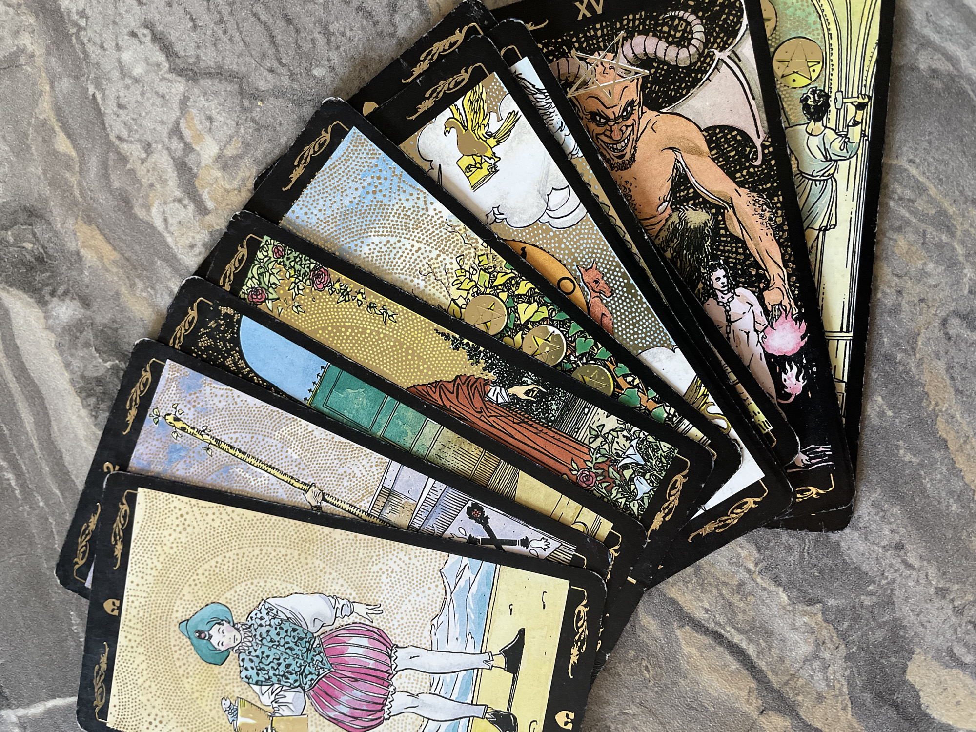 An overhead shot depicts a spread of tarot cards fanned out on a gray marble surface. The card furthest to the right is marked "XV" and displays a depiction of the Devil. Progressing left, the card second from the right shows a person with a hammer next to vertical lines. The third card from the right depicts a golden eagle. The remaining cards are partially obscured, but visible elements include gold coins, green towers, and a person in a pink and blue outfit. The environment appears to be indoors due to the lighting and the presence of the marble surface. The image does not show Izhevsk, Russia.