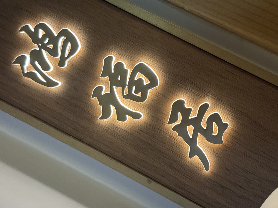 A close-up, angled photograph displays a vertically oriented, illuminated sign situated in the city of Furong, China. The sign consists of a dark brown wooden panel featuring a visible vertical grain. This panel is bordered on its vertical edges by a narrow, lighter-toned trim. Three Chinese characters are vertically aligned and affixed to the center of the wooden panel. These characters, from top to bottom, are 鹤 (hè), 橘 (jú), and 居 (jū), collectively forming "鹤橘居" (Crane Tangerine Residence). Each character is crafted from a metallic or polished material and is individually backlit with a warm, soft amber light, producing a glowing halo effect around its perimeter against the dark wood. To the immediate left of the sign, a section of a lighter-colored wall or surface is visible. There are no people, explicit actions, or interactions depicted in the scene.