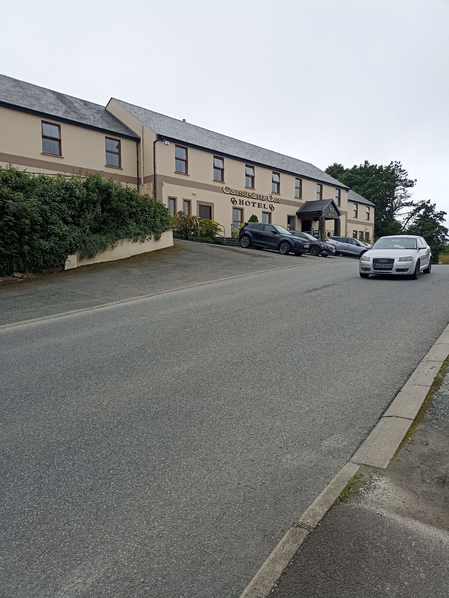 The image captures a frontal view of the Castles of Oir Hotel in Annagry West, Ireland, on an overcast day. The hotel is a two-story building with a beige facade, a dark grey tiled roof, and numerous brown-framed windows. A prominent sign above the entrance reads "CASTLES OF OIR HOTEL". Green hedges line the front of the hotel, partially obscuring the lower level. In front of the hotel, parked on a sloped asphalt area, are several cars: a dark grey SUV on the left, a black sedan behind it, and a silver Audi A3 in the foreground, which is in motion, approaching the viewer. The road in front of the hotel is also asphalt and slopes downwards. The sky is a uniform grey, indicating cloudy weather. A concrete curb and gutter are visible along the right side of the road.