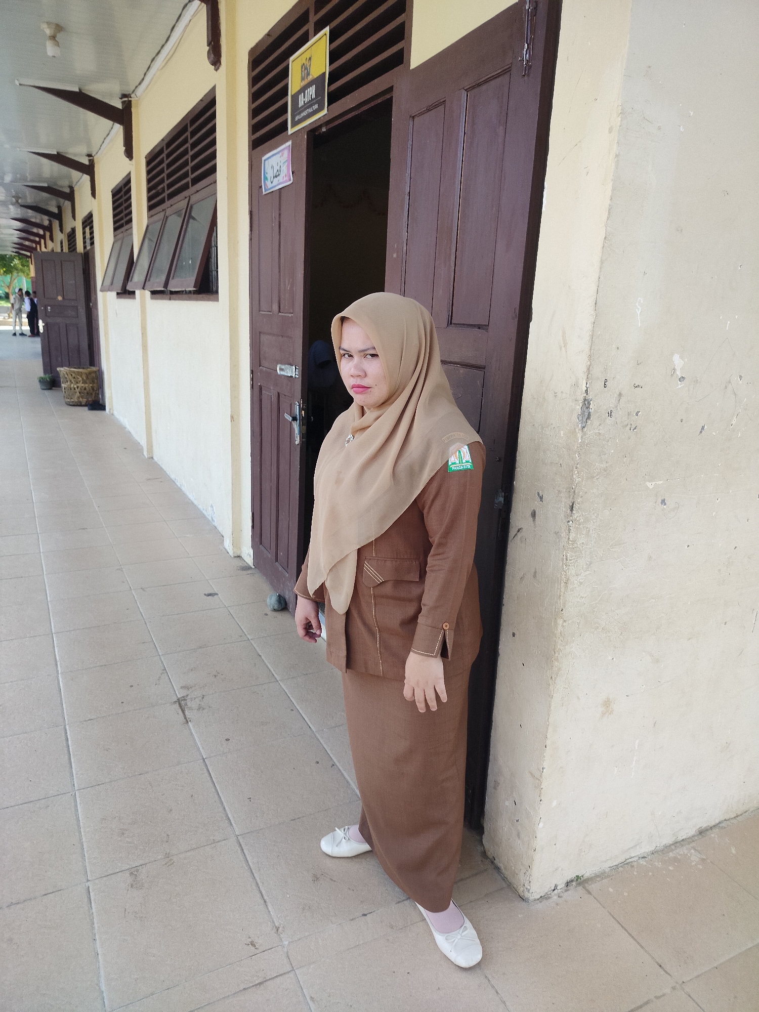A woman wearing a brown hijab and a matching brown uniform stands on a tiled walkway in front of a building. The uniform has a patch on the left sleeve with a green and white logo. She is looking to her right, with a neutral expression. Behind her are two brown wooden doors, one partially open, revealing a dark interior. Above the open doorway, a sign with yellow and black lettering is visible. The building's exterior wall is a pale yellow color and appears worn. To the left of the woman, a long covered walkway extends, with several open windows with brown frames. The ground is tiled with light brown squares. In the distance, to the left, other people can be seen walking in the shade of the walkway. A woven basket is placed near one of the doors. The scene suggests an outdoor school or office environment in a warm climate.