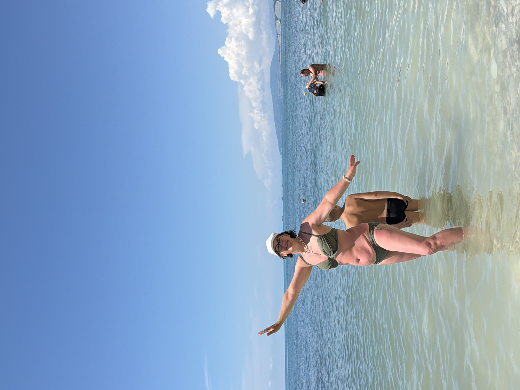 A woman in a green bikini and white baseball cap stands in shallow, clear water at a beach, with her arms outstretched to her sides. Behind her, a boy in black swim trunks stands in the water. In the background, several people are in the ocean, some swimming and others standing. The sky is clear blue with a few white clouds on the horizon. The scene is the coast of Divnomorskoe, Russia, known for its pebble beaches and clear waters.