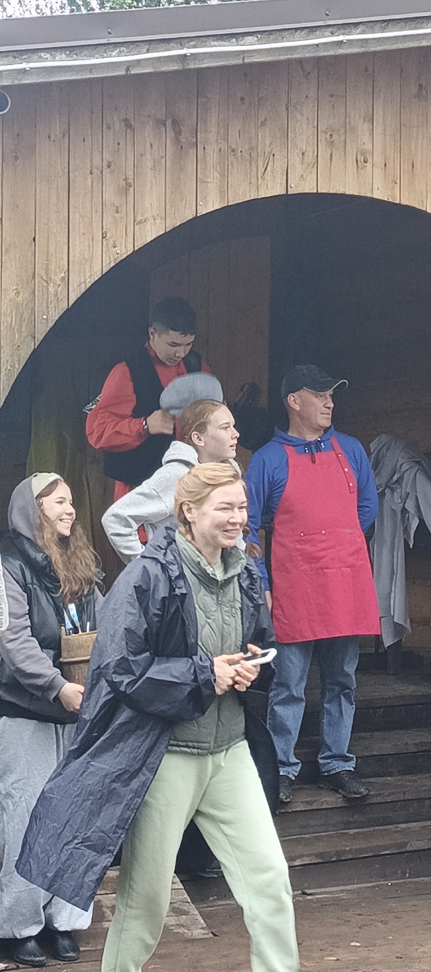 A group of people stand on wooden steps in front of a wooden structure with an arched opening. The structure appears to be a building or an entranceway. The people are dressed casually, some in raincoats, and one man wears a red apron. The lighting suggests an overcast day. The location is identified as Старые Быги, Russia.