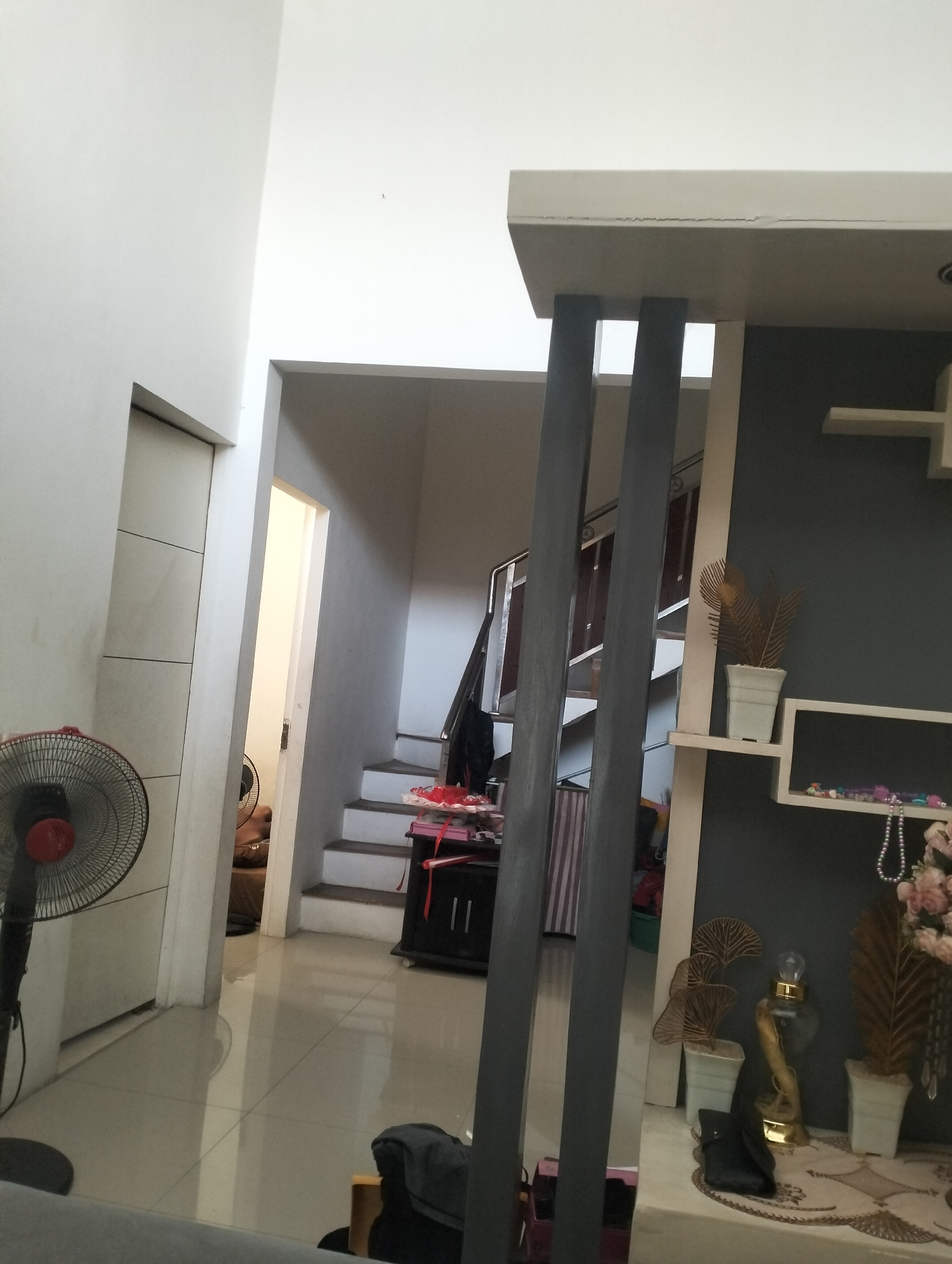 This is an indoor scene captured from a medium-low angle, showing a contemporary living space with an open-plan layout. The foreground features a tall, oscillating fan on the left, partially obscuring a white, vertically segmented wall. To the right of the fan, a doorway leads into a dimly lit hallway or room, with another fan visible in the distance.
The mid-ground is dominated by a staircase that curves to the right, with white steps and dark railings. A dark cabinet is positioned at the base of the stairs, adorned with red and white decorative items. A partition with vertical dark grey wooden elements separates this area from the right side of the image.
The right side of the image showcases a wall with a dark grey finish, accented by a white shelving unit. This shelf holds decorative items, including a white pot with a dried plant, a glass bottle with a gold stopper, and another dried plant in a white pot. Below this, a white rectangular shelf displays beaded necklaces and pink flowers in a vase. The overall aesthetic is modern and minimalist, with a neutral color palette. The flooring appears to be polished light-colored tiles. The lighting is diffused, suggesting an interior setting during the daytime, possibly with natural light filtering in from unseen sources.