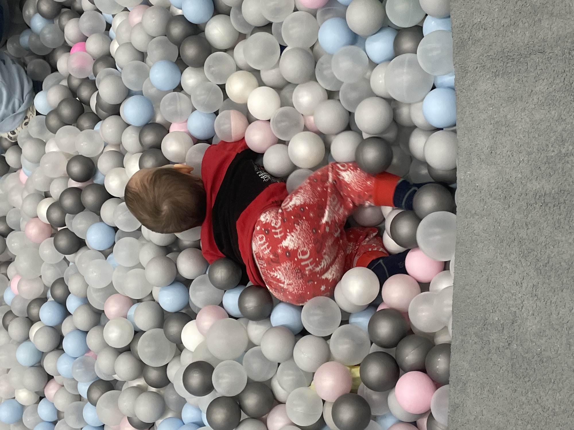 A child in red patterned pajamas and a black top lies curled amidst a ball pit filled with gray, white, pink, and blue plastic balls. The ball pit appears to be in a corner, bordered by a gray wall. The location is Perm, Russia.