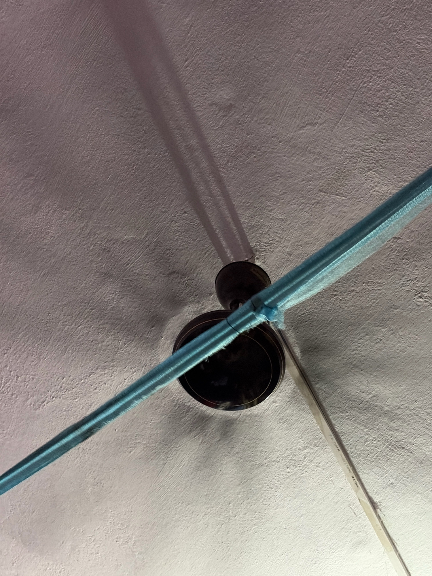 A low-angle shot focuses on a ceiling fan in Jamshedpur, India. The fan, with dark brown blades, is partially obscured by a bright blue mesh material wrapped around one of its blades. Shadows from the fan blades are cast onto the textured white ceiling. A thin white cable runs diagonally across the lower right portion of the image, also casting a faint shadow. The lighting appears to be artificial, creating a slightly warm and diffused glow. The environment suggests an indoor setting, likely a room.