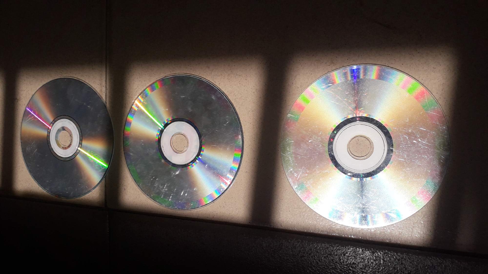 Three CDs are arranged in a row on a light-colored, tiled surface. A shadow, likely from a barred window or similar structure, falls across the scene, creating areas of light and dark. The CDs are positioned close together, with the smallest on the left and the largest on the right. The surfaces of the CDs are reflective and show a rainbow-like diffraction pattern. There are no people visible. The location is Nkpor, Nigeria.