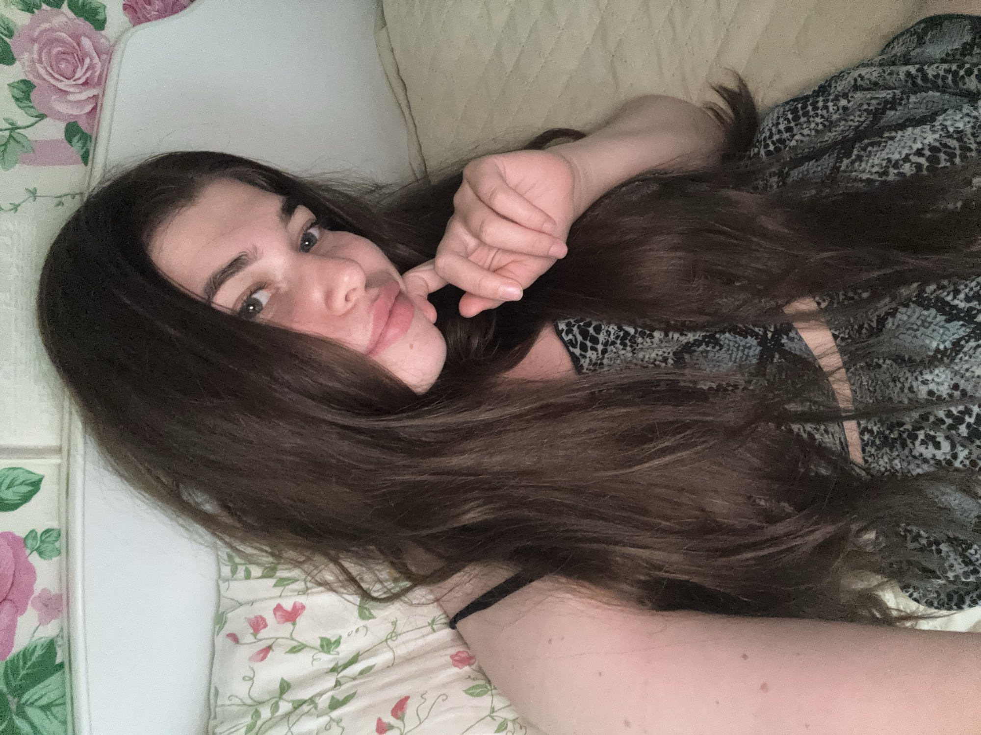In the city of Мещёры, Russia, a young woman with long, dark brown hair lies on a bed. She is wearing a black and gray snakeskin-patterned top and a black bra strap is visible. Her right hand is raised, with her fingers resting on her chin and mouth. The background consists of floral-patterned pillows and a textured bedspread.