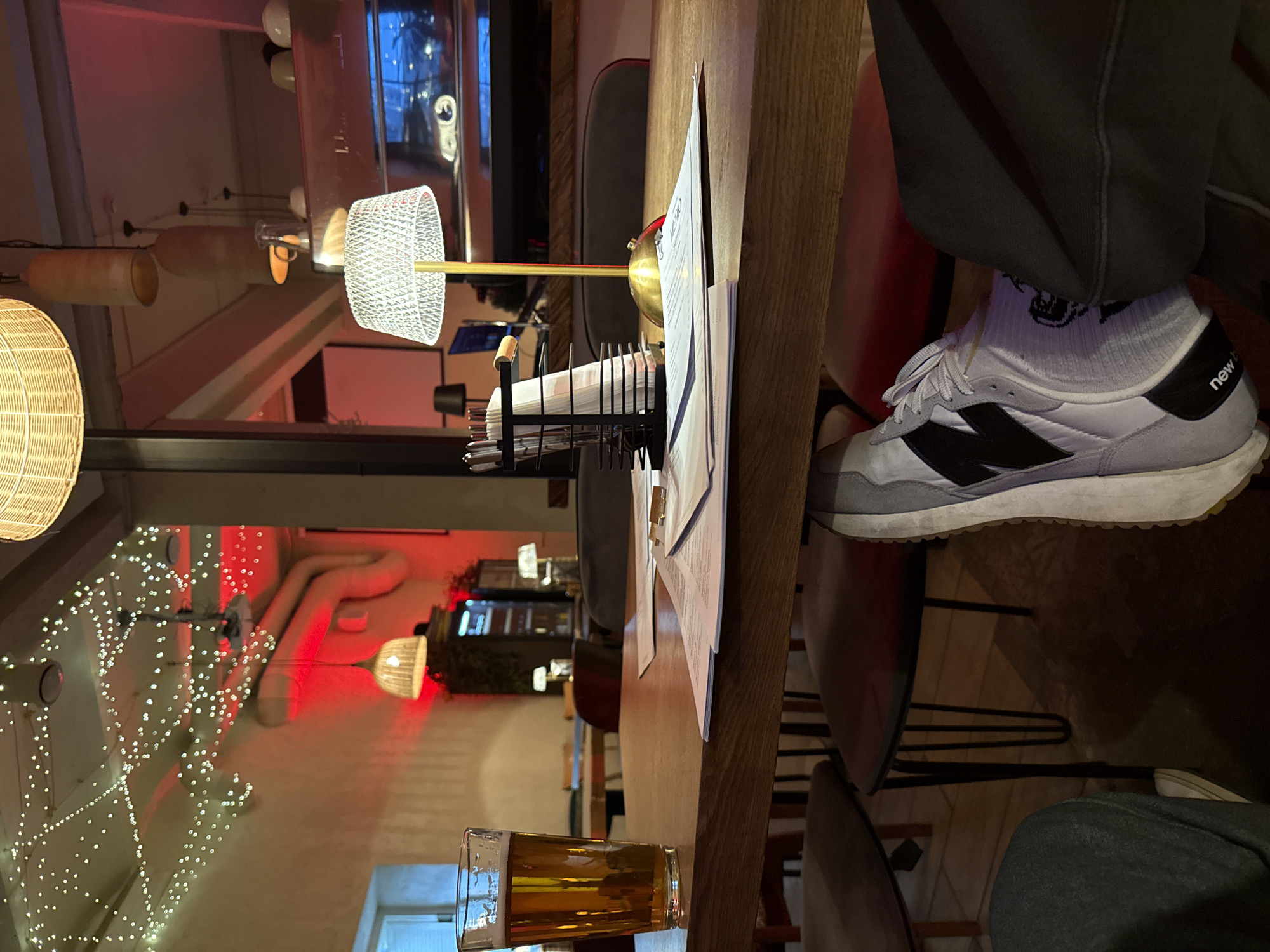 The image shows a table in what appears to be a restaurant or bar, likely in Moscow, Russia, given the Cyrillic script visible on the menu. A glass of beer sits on the wooden table, near a metal napkin holder and a menu. A person wearing grey sweatpants and New Balance sneakers has one foot resting on the chair next to the table. The background features a festive atmosphere with decorative lights, potentially for a holiday season. There are other chairs and tables, along with lighting fixtures visible.