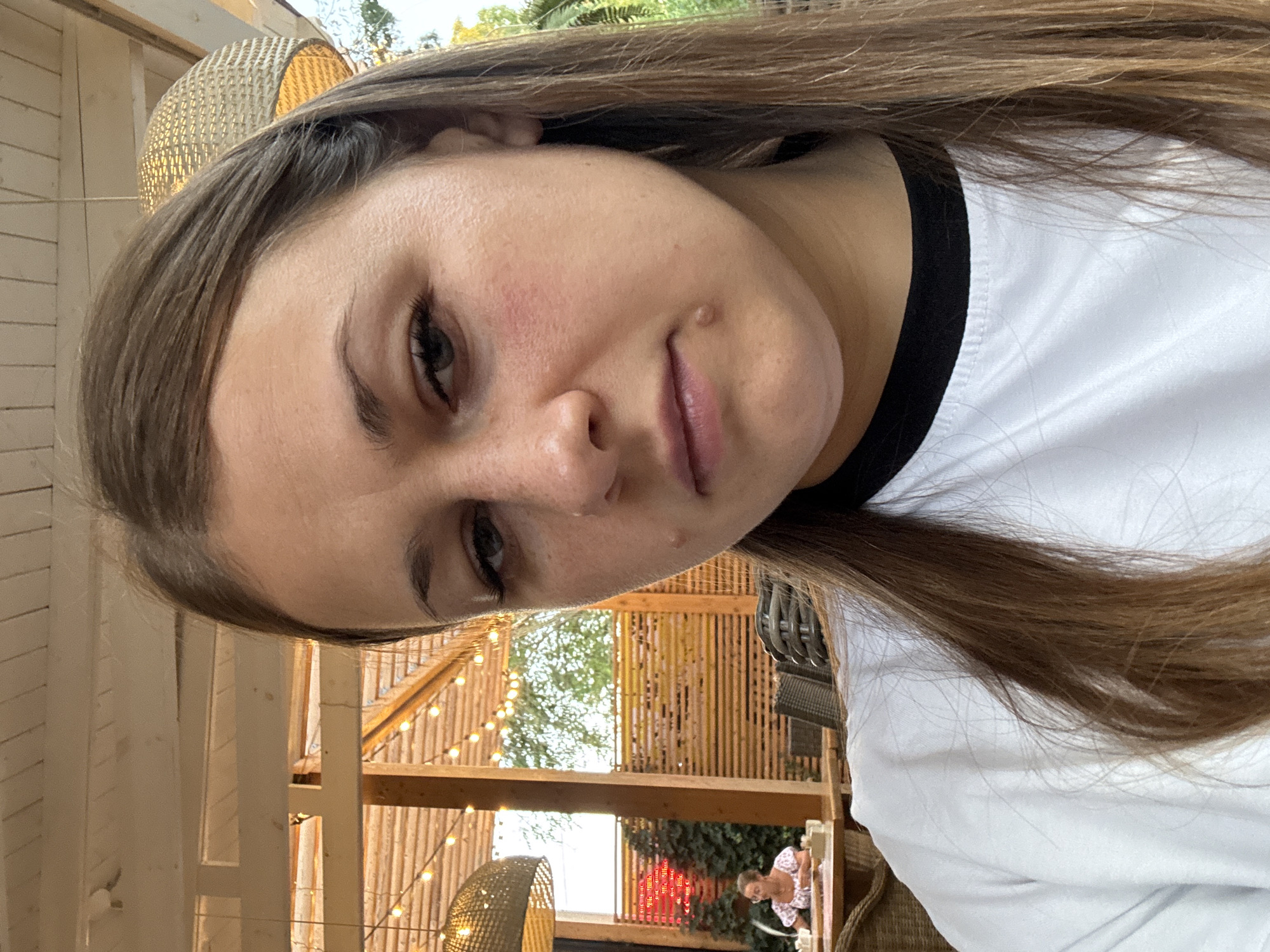 A young woman with long, brown hair takes a selfie in what appears to be an outdoor cafe in Yevpatoriya, Ukraine. She is wearing a white t-shirt with a black neckline. Her expression is neutral, and she is looking directly at the camera. In the background, wooden structures and string lights are visible, suggesting an evening or late afternoon setting. A woman is seated at a table in the distance, and there are chairs and tables arranged throughout the outdoor space. Lush green foliage can be seen behind the wooden fence. The overall environment suggests a relaxed, casual atmosphere.
