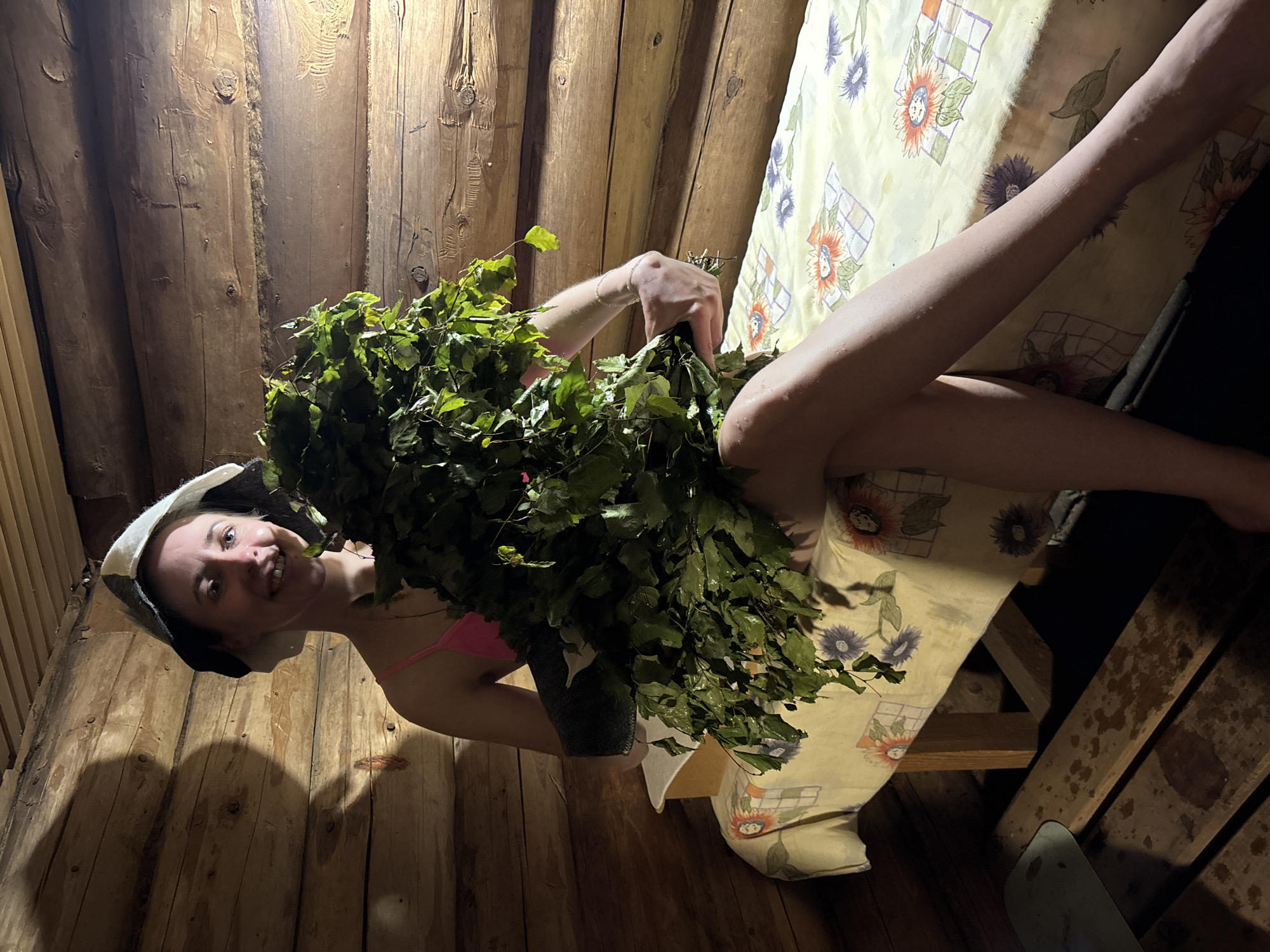 A woman wearing a sauna hat and pink swimwear sits in a wooden sauna in Chaikovsky, Russia. She is holding a large bundle of leafy green branches. The interior of the sauna is lined with wooden planks. The woman is positioned beside a bench covered with a patterned light yellow cloth.