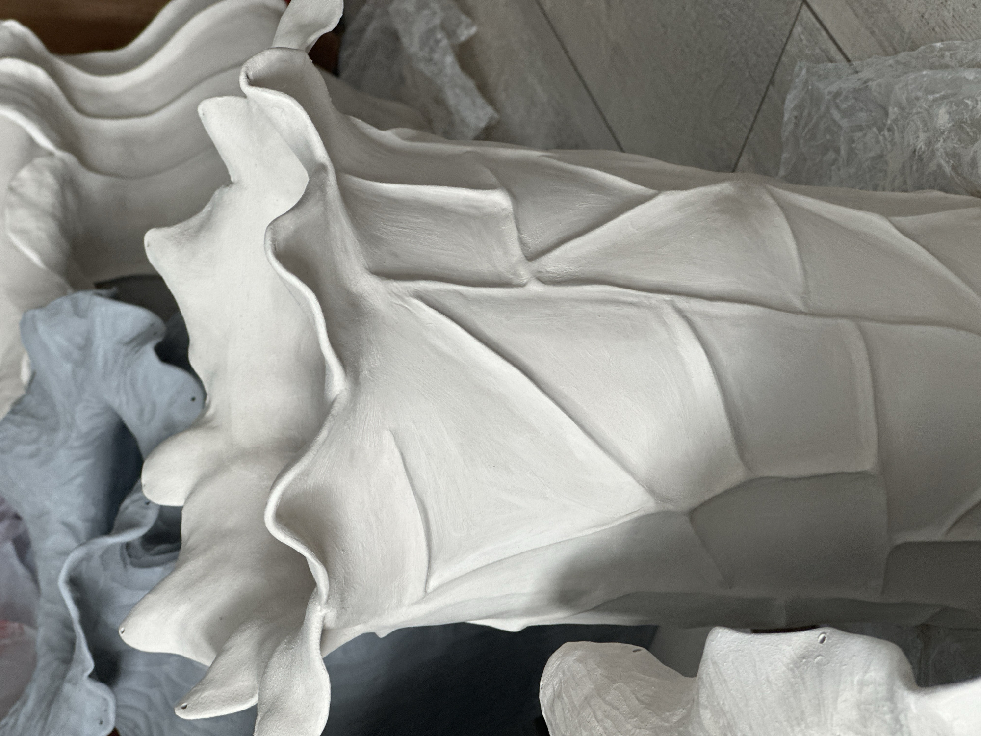 The image shows a close-up of a white, petal-shaped ceramic vase with a textured, faceted surface. The vase has a ruffled, open top and appears to be unfinished or unglazed. In the background, other similar ceramic pieces, some in a pale blue color, are visible. The objects are placed on a light-colored tiled floor, with some appearing to be wrapped in plastic. The setting is indoors, and based on the objects, it is likely a studio or workshop. There is no indication of a specific location within the city of Nevinnomyssk, Russia.