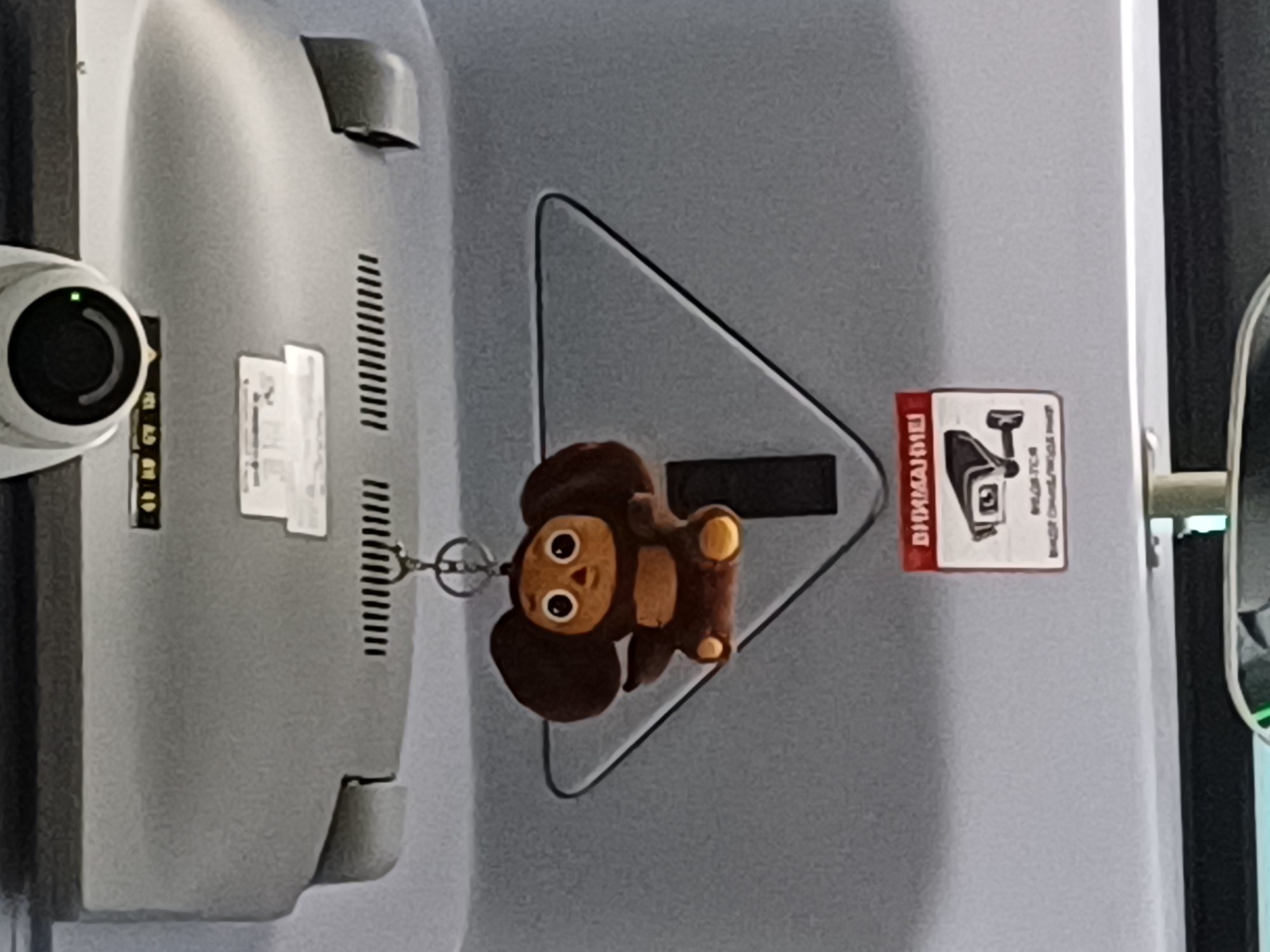 A plush toy keychain of a cartoon character, Cheburashka, is hanging from a metal ring attached to the ceiling of a vehicle. The toy is brown with large ears and eyes. Below the toy, a red and white sign with an image of a security camera and Russian text is affixed to the ceiling. The text on the sign reads "ВНИМАНИЕ!" which translates to "ATTENTION!". The ceiling of the vehicle appears to be made of grey plastic, and a security camera is visible at the top of the image, positioned above the toy and the sign. The background shows the interior of the vehicle, suggesting it could be a bus or a van. The image is taken from a low angle looking upwards.