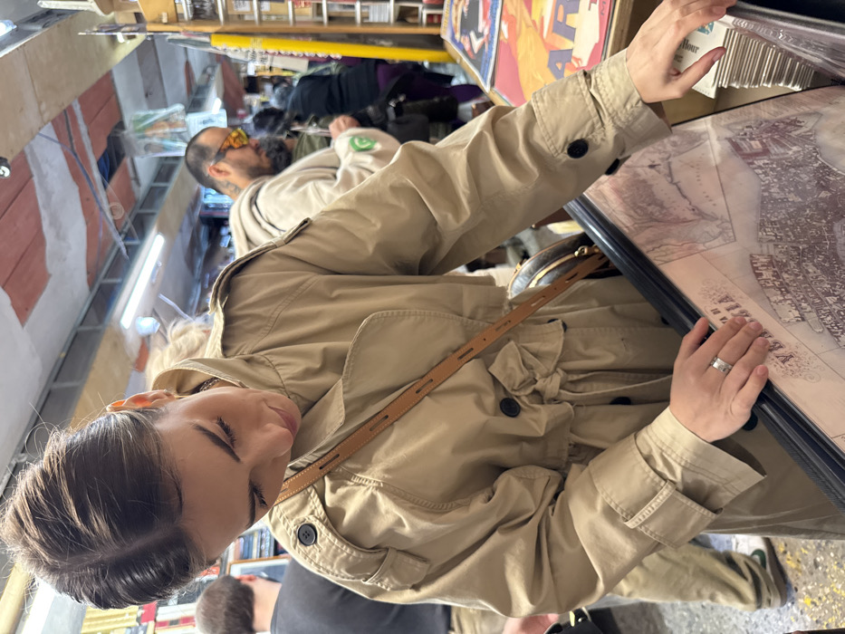 The image displays an indoor market stall in Venice, Italy. A young adult female, with dark hair styled in a bun, is the primary subject, viewed from the upper torso down, leaning forward and looking intently downwards. She wears a tan double-breasted trench coat with a belted waist and a light brown leather cross-body bag. A silver-colored ring is on her left ring finger. Her hands are on a large, framed or mounted print, identified by the text "VENEZIA" on the visible map or diagram. To her immediate right, a stack of similar prints or flat items is partially visible.
In the background, other individuals are present in varying states of focus. To the far left, the back of a man wearing a dark shirt is visible. To the right of the main subject, a man with short dark hair, neck tattoos, and orange-tinted sports sunglasses is seen from the chest up, facing away and slightly to the right. He wears a light-colored hooded sweatshirt with a green symbol on the left sleeve. Additional out-of-focus figures, including one wearing purple pants, are discernible further into the crowded market. The environment features overhead fluorescent strip lighting, exposed ceiling structures, and shelves stocked with various items, consistent with an antique or specialty market stall.