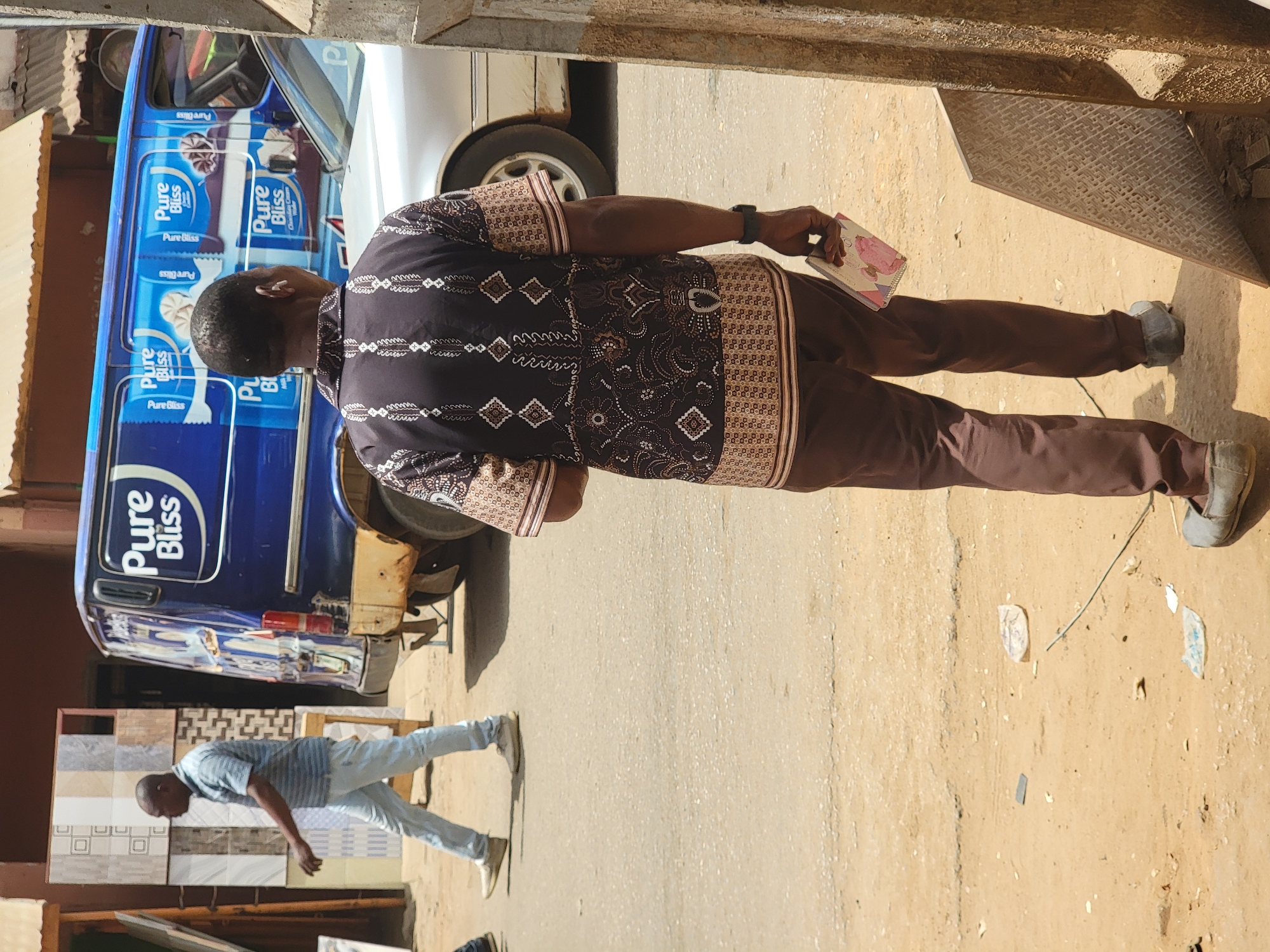 The image shows a man in a patterned shirt and brown pants walking on a dirt road in Maiburiji, Nigeria. He is carrying a book or notebook. A blue vehicle with "Pure Bliss" branding is parked nearby. Another man is walking in the background, near some tiles. The environment appears to be an urban or peri-urban area. There are scraps of litter on the ground.