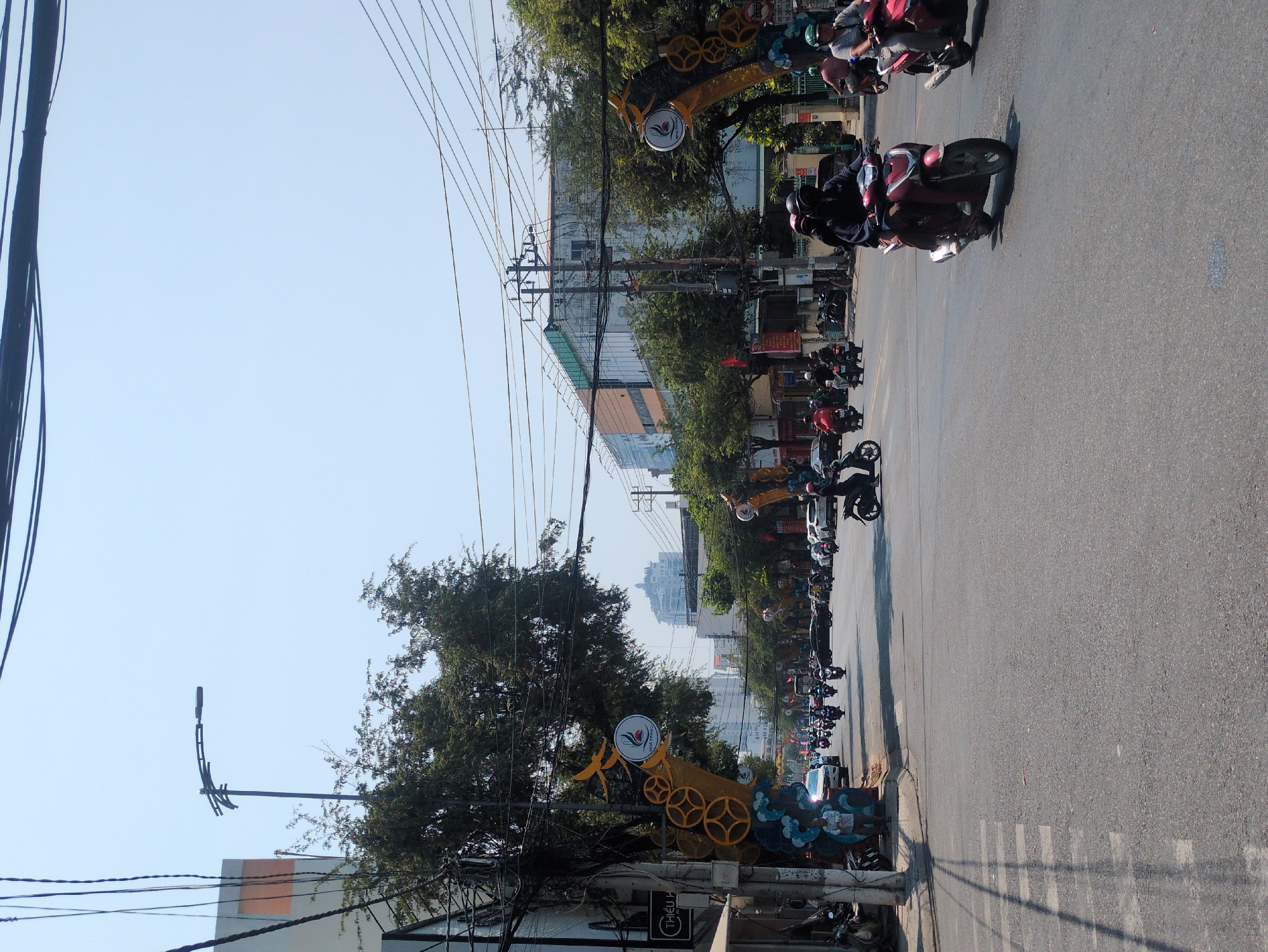 The photograph shows a street scene in Nha Trang, Vietnam. A line of motorbikes is heading down the right side of the road. The street is asphalt and appears dry. Overhead power lines crisscross the sky, partially obscuring buildings in the background. Street signs and trees line the road, with a large decorative golden sign at the roadside. The sky is clear and light blue.