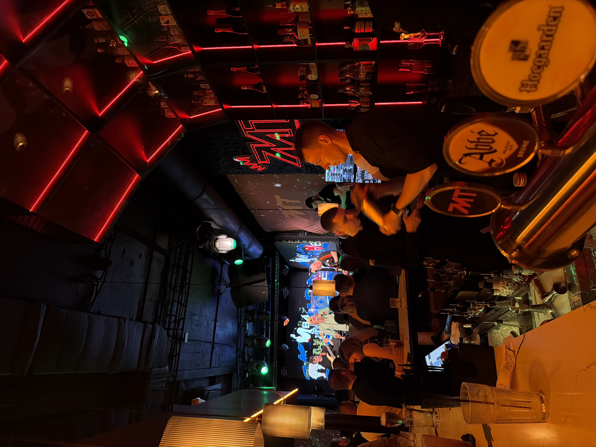 The image depicts a dimly lit bar with a retro, possibly industrial aesthetic. Red neon lights trace geometric patterns on the ceiling, reflecting on polished surfaces and illuminating rows of bottles behind the bar. Several people are visible, some standing behind the bar and others seated or standing in the background, suggesting a social gathering or a busy evening service. One person in the foreground, a man with short hair wearing a black t-shirt, appears to be tending to the bar or interacting with customers.
Prominently displayed on the bar are three beer taps, each with a distinctive emblem. The top tap features a circular logo with the text "Hoegaarden." Below it, another tap displays a logo with the word "Abbé." The third tap has a logo with "MT." These brands indicate the types of beer likely available at the establishment.
In the background, a large screen displays a colorful, animated scene with cartoonish characters, adding a playful or entertainment element to the bar's atmosphere. The overall mood seems lively and relaxed, typical of a bar environment in the evening. The lighting creates a warm and somewhat moody ambiance. No specific weather conditions are discernible. The visible text on the tap logos provides direct clues about the products offered. The location context of Khabarovsk, Russia, suggests this is a bar within that city.
