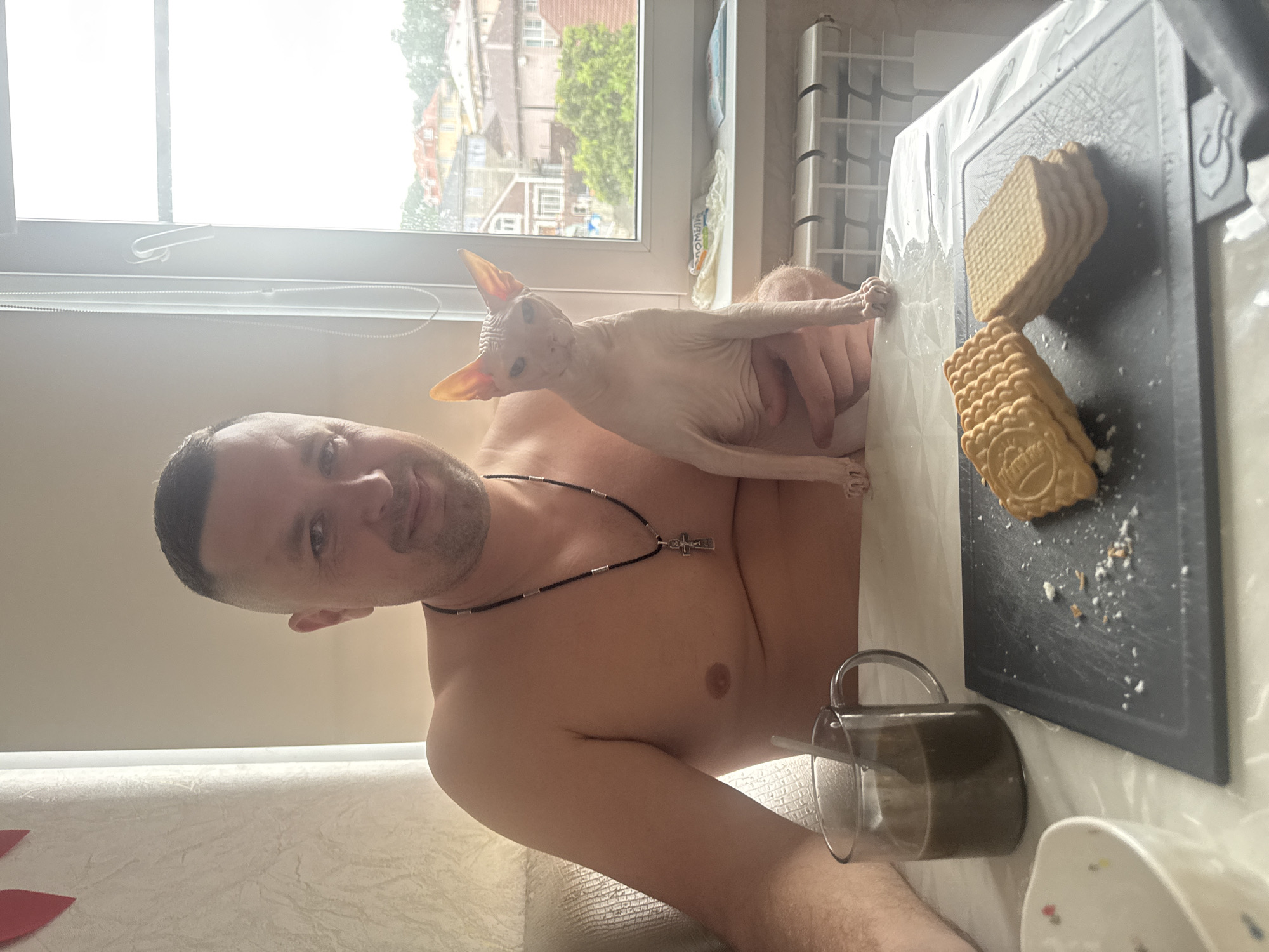 A shirtless man with short dark hair is holding a Sphynx cat. The man is wearing a necklace with a cross pendant. The cat is resting on the man's chest and extending its paws towards a stack of sandwich crackers on a black cutting board. A clear mug with dark liquid is on the table next to the cutting board. A bowl is partially visible in the lower right corner. A window in the background shows a view of buildings and trees, suggesting a residential area. Based on the presence of buildings and trees in the background and the description of the location, the scene could be situated in Vesyoloye, Russia, but this is only possible to determine.