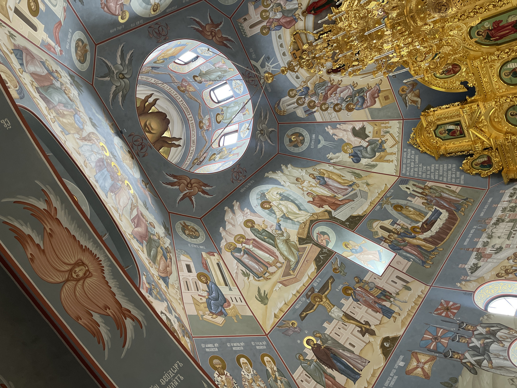 The image shows the interior of a Russian Orthodox church ceiling, likely in Khimki, Russia. The ceiling is richly decorated with religious iconography and frescoes, depicting various saints and biblical scenes. The color palette is dominated by blues, golds, and earth tones. A central oculus provides natural light. A large, ornate gold chandelier hangs partially in the frame. The perspective is from a tilted angle, looking upwards. Inscriptions in Cyrillic script are visible along the lower portions of the painted sections.