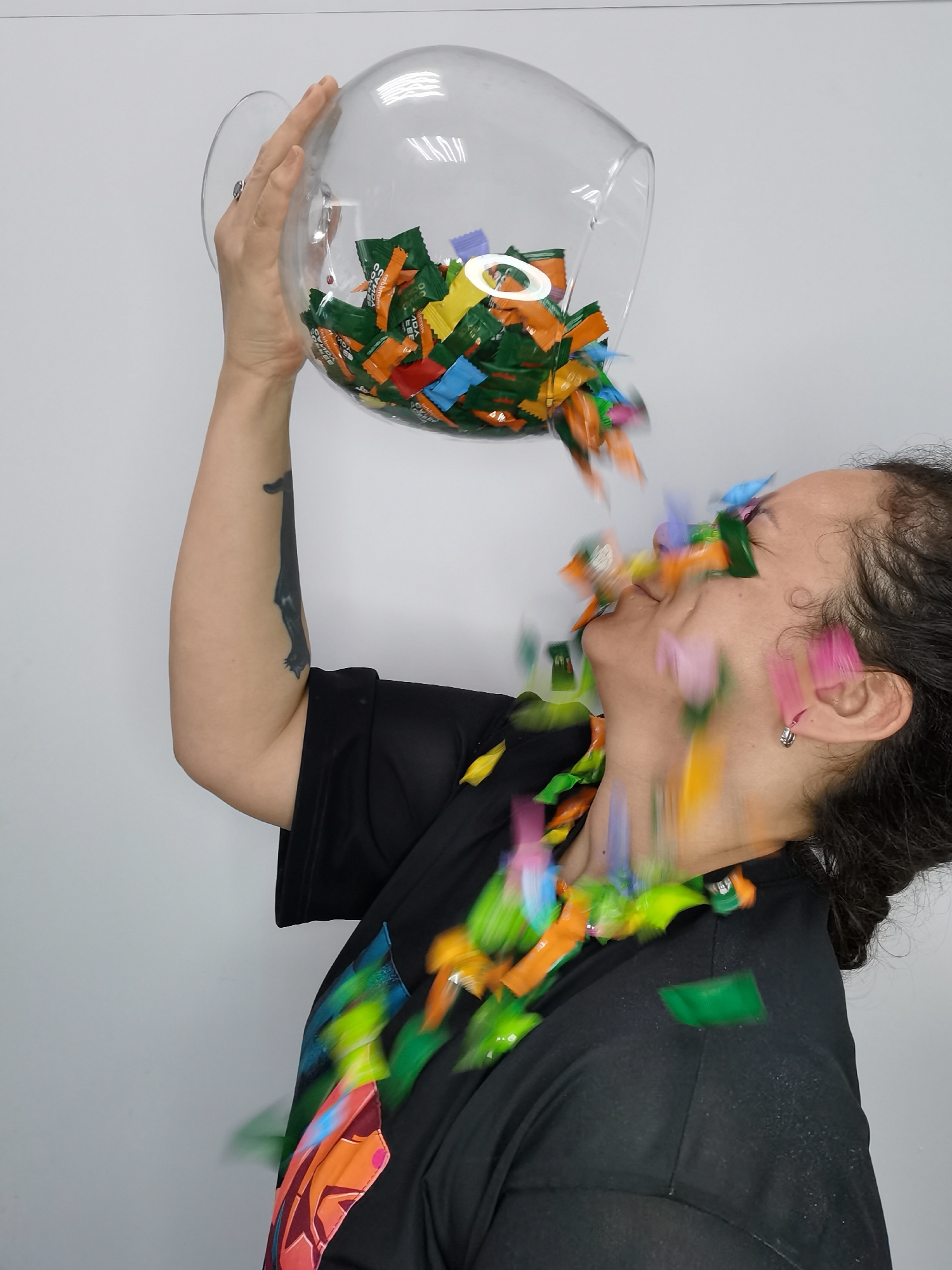 A person is standing against a white wall, holding a large, clear glass bowl filled with wrapped candies above their head and tipping it to pour the candy over their face and into their open mouth. The individual has dark hair pulled back, fair skin, and is wearing a black shirt with colorful designs on the front. A black cat tattoo is visible on their left arm. The candy wrappers are a variety of colors, including green, orange, yellow, and blue. The person is smiling. The location is likely an indoor setting with a plain, white wall. The city of Chaikovsky, Russia is not determinable from the image.