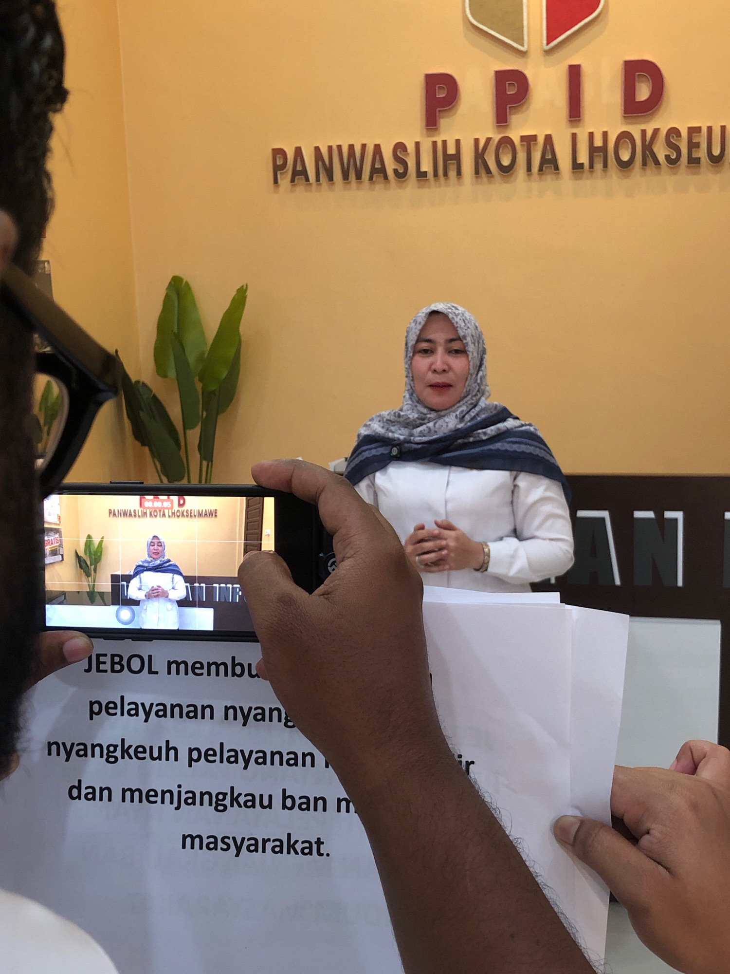 A medium shot captures a woman speaking in front of a yellow wall adorned with large, dark lettering that spells "PPID PANWASLIH KOTA LHOKSEU". The woman, wearing a white collared shirt and a grey and blue patterned headscarf, stands with her hands clasped in front of her. A smartphone is being held in the foreground, pointing towards the woman and displaying her image on its screen. The phone's screen also shows the text "PANWASLIH KOTA LHOKSEUMAWE". In the lower part of the image, a white paper with dark text is held by a person whose arm is visible on the right. The text on the paper is partially obscured but reads, "JEBOL memb," followed by lines of Indonesian text. The background includes a potted plant and a sign with the word "GRATIS" on it. The overall scene appears to be an indoor setting, possibly an office or public building in Lhokseumawe, Indonesia.