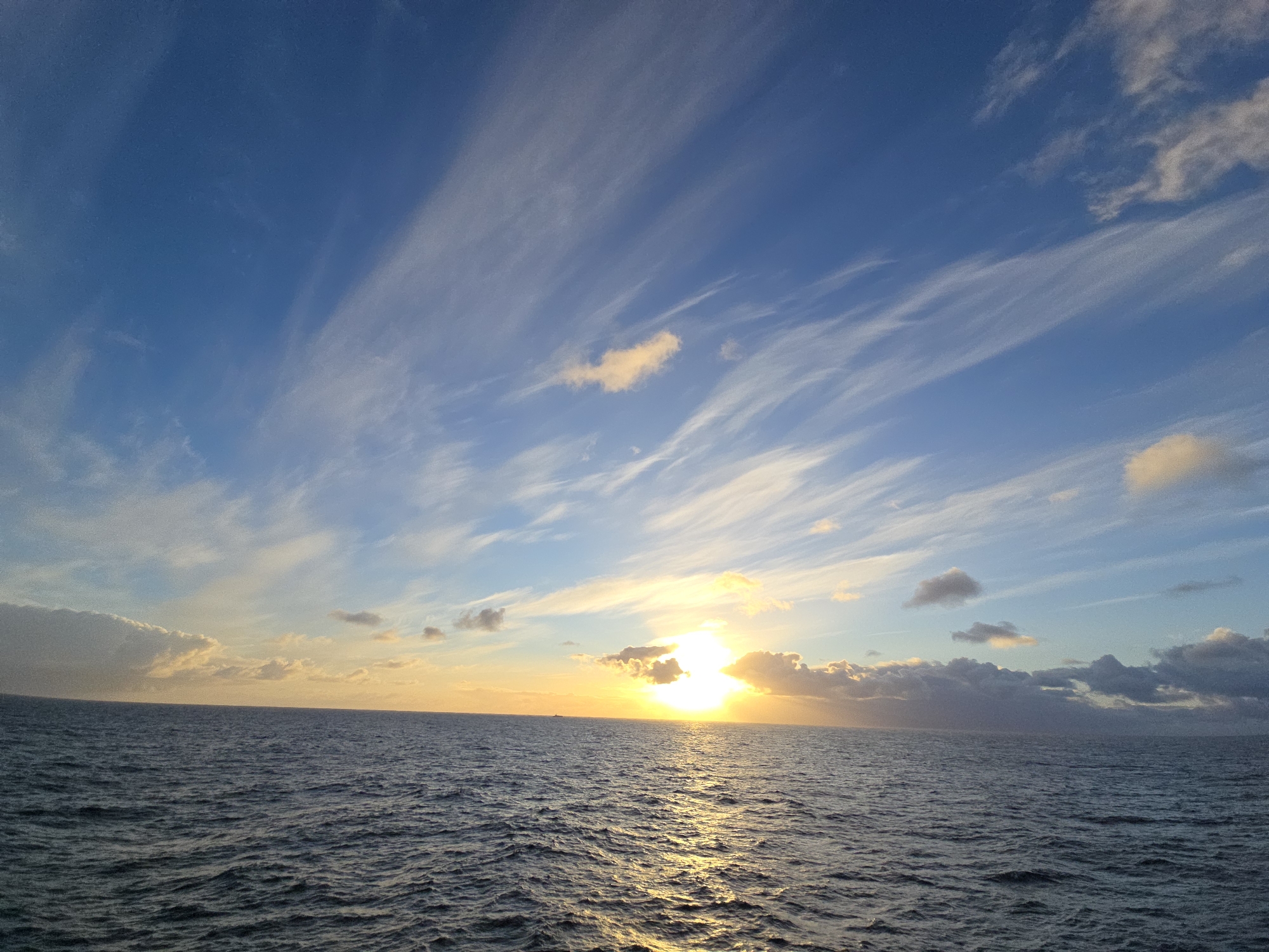 The image shows a sunset over the ocean. The sky is blue with wispy clouds. The sun is setting on the horizon, with light reflecting on the water. The location is at sea and the city is Unknown, Unknown.