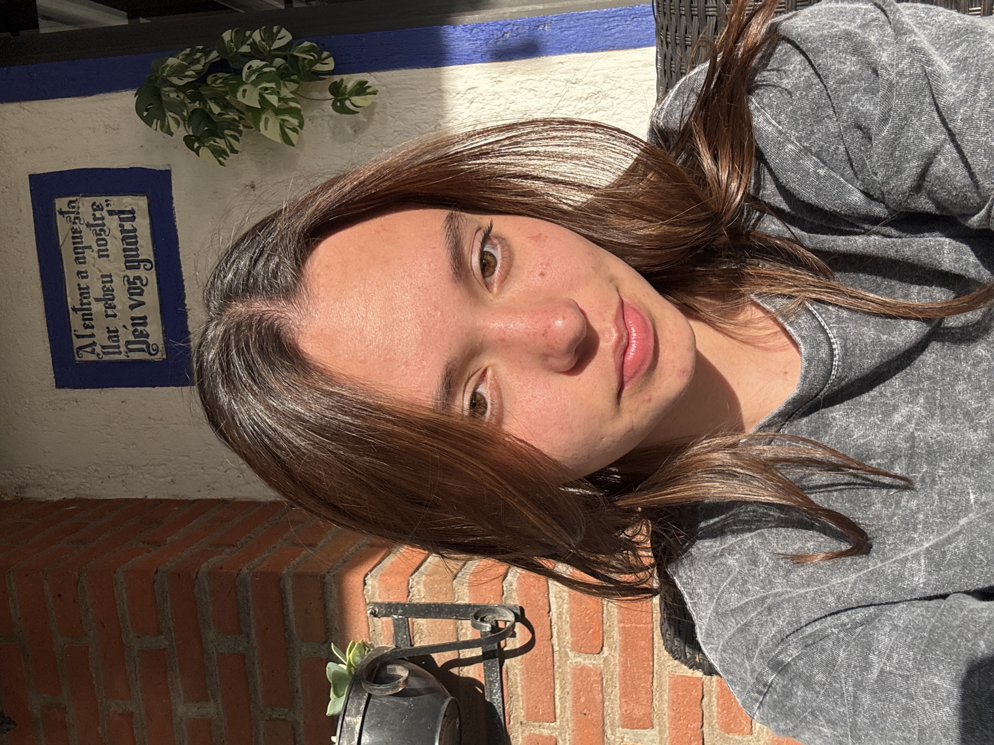 A young woman with brown hair and a gray shirt poses for a selfie. She is turned slightly to the side, with her face angled toward the camera. A portion of a brick wall is visible in the background, with a decorative metal piece and a small plant attached. Above the brick wall, there is a white wall with a blue stripe at the top. A plaque with text is also visible on the white wall, along with a small plant. The location is in Puebla, Mexico.