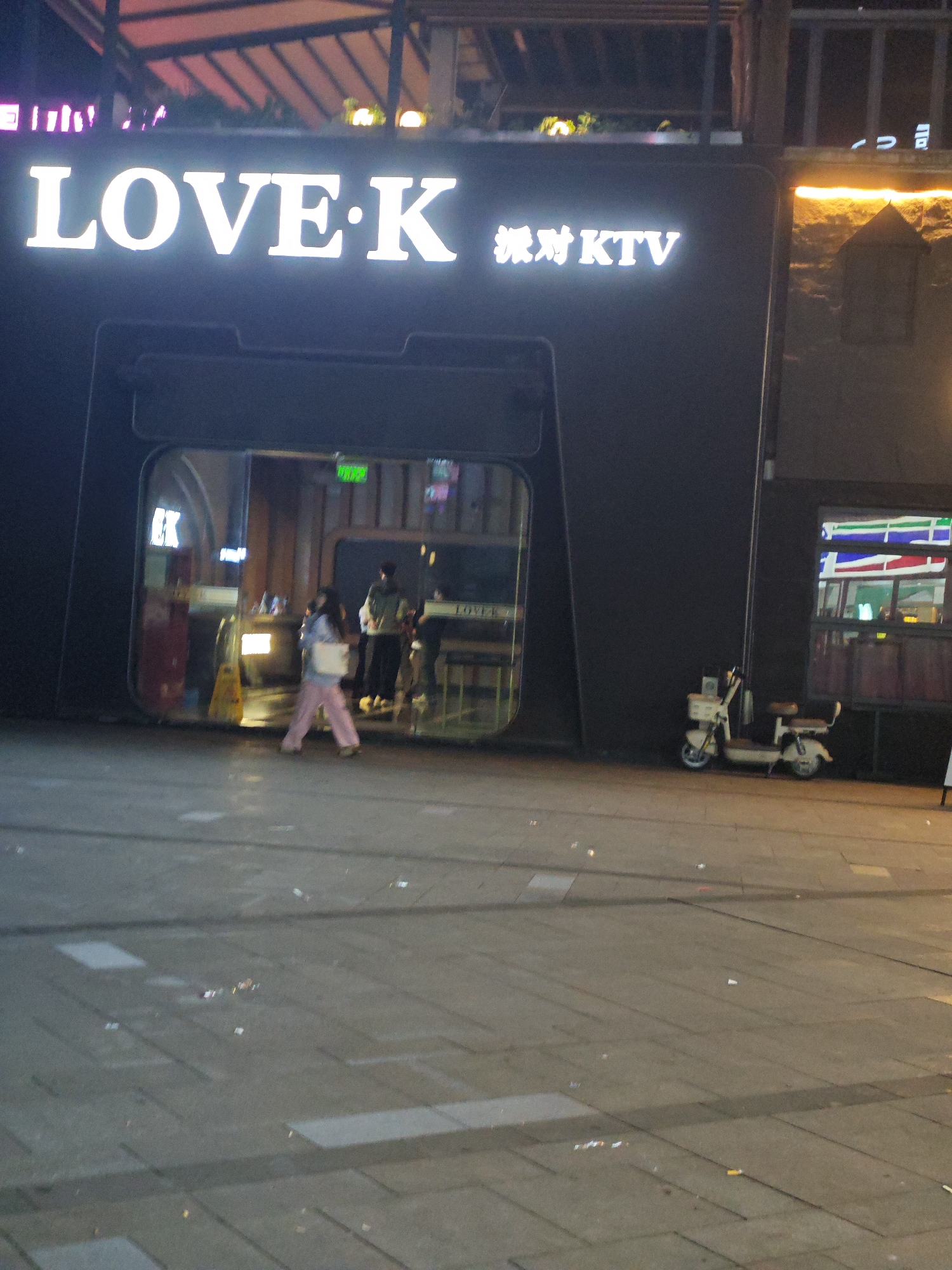 The image captures an exterior view of "LOVE-K 派对KTV" (Party KTV) located in the Chongqing High-tech Zone, China. The building is dark, with bright white signage illuminating the night. An entrance with a glass facade reveals individuals inside. A woman with pink trousers is seen walking out. A parked scooter is visible to the right of the entrance, against the building's dark exterior. The surrounding pavement is made of large stone slabs and is littered with debris.