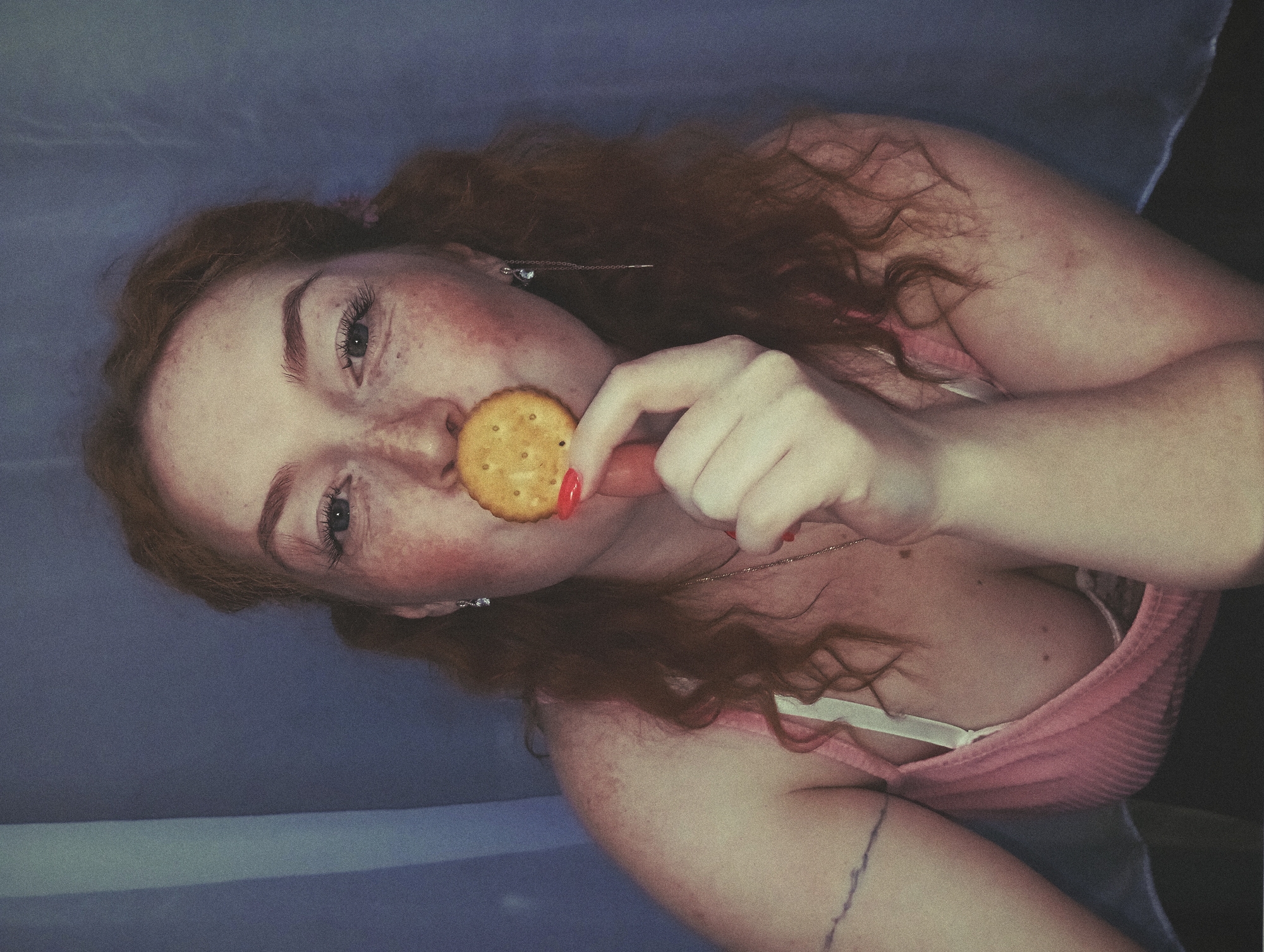 The image shows a young woman with red hair and freckles holding a round cracker to her mouth with her right hand. Her fingernails are painted red. She is wearing a pink tank top. The background appears to be a light blue or purple fabric or wall. The location is not discernible.