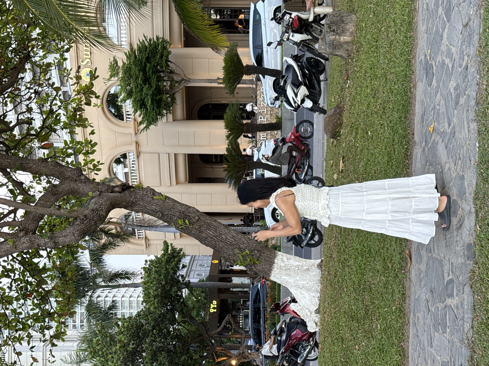 A woman in a white tiered dress and black sandals stands on a stone pathway, holding a smartphone to take a picture. She is facing away from the camera and looking towards a street scene. Behind her, a large tree with white-painted bark stands in a grassy area. The street features several parked motorcycles and cars, as well as palm trees and a light-colored building with arched windows and columns. The building has a sign with a yellow sun logo and the word "Sun" visible. The street is lined with shops and businesses, with people visible on the sidewalks and in some of the establishments. The environment appears to be a busy urban area in Nha Trang Ward, Vietnam, likely a commercial district near the coast.