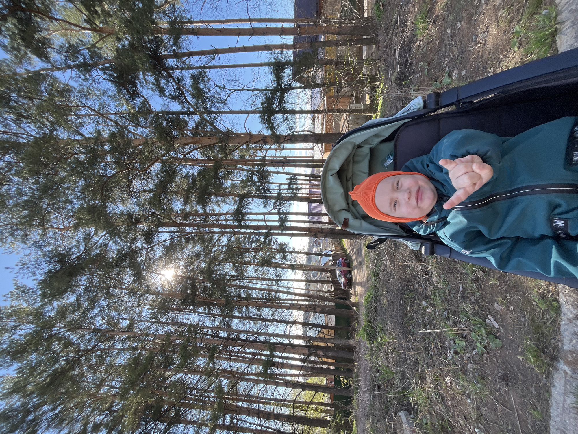 A child wearing a green jacket and an orange hat sits in a stroller. The child is making a hand gesture. The stroller is on a path lined with tall trees. The location appears to be in a wooded area near a body of water. There are cars and buildings visible through the trees. Given the prompt, it's plausible that this is Chaikovsky, Russia.