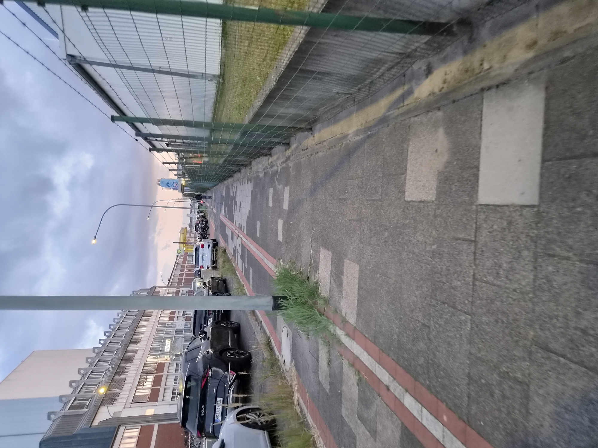 A perspective shot of a sidewalk running alongside a fence and a road with parked cars, likely in Bremerhaven, Germany. The sidewalk is made of concrete pavers with red and white painted lines, and grass grows through some of the cracks. Parked cars, including a black Kia Sportage with a German license plate "HB:A 1603E," are visible on the right side of the road. A large, modern building with many windows is on the left side of the image. A barbed-wire fence runs along the right side of the sidewalk, separating it from a grassy area. The sky is overcast with muted colors, suggesting it is late afternoon or early evening.