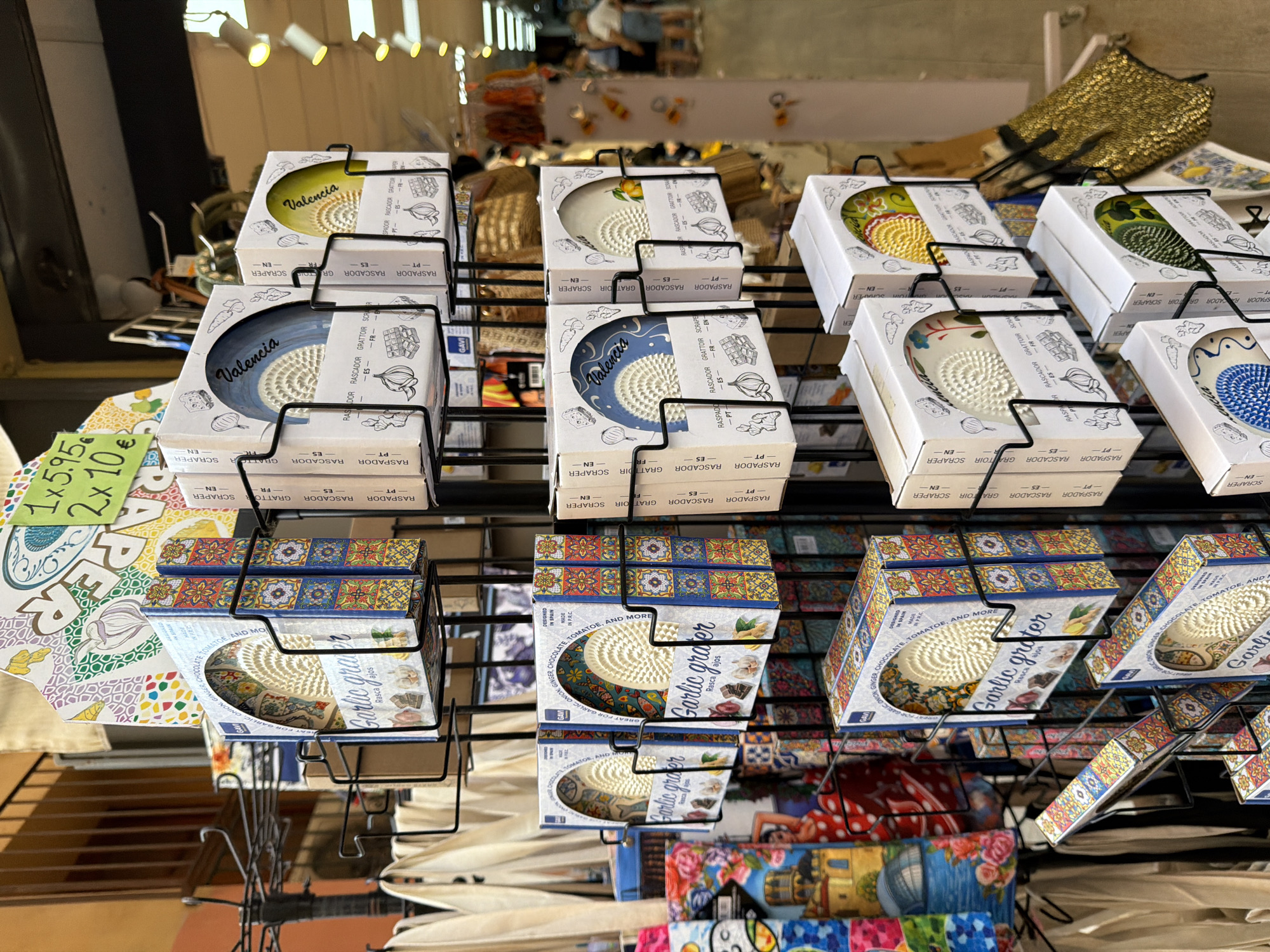 The image shows a close-up of a retail display stand filled with garlic graters packaged in colorful boxes. The stand is filled with multiple levels of these graters, with some boxes facing forward and others sideways. The boxes feature decorative patterns, often with a Mediterranean or Spanish tile motif, and text indicating "Garlic grater" and "Valencia." Some boxes have cut-out windows revealing the white ceramic grater inside. The background suggests an indoor market or shop setting, with other products visible on shelves and the floor, including fabric bags, textiles with colorful prints, and some indistinct merchandise. Lighting appears to be artificial, coming from overhead fixtures. In the blurred background, several people are visible, appearing to be shoppers. The scene is brightly lit and appears to be a point of sale in a souvenir or specialty food shop.