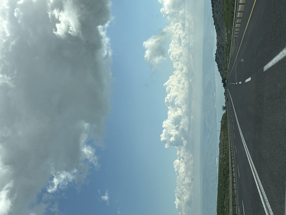 The image displays a two-lane asphalt highway descending towards the Sea of Galilee (Kinneret), observed from an elevated position in the vicinity of Safed, Israel. The sky occupies the upper two-thirds of the frame, featuring a large, dark grey cloud mass in the upper left quadrant, transitioning to a clear blue sky with scattered white cumulus clouds along the distant horizon. The highway, marked with continuous white lines on its outer edges and a double solid white line separating the two directions of travel in the foreground, extends into the distance, curving slightly left. Metal guardrails line both sides of the road. To the left, a green grassy slope with visible rocks borders the highway. To the right, the terrain is rocky and uneven with sparse green vegetation. Two vehicles are discernible in the distant section of the highway: a dark-colored car traveling away from the viewer on the left lane, and a lighter-colored car traveling towards the viewer on the right lane. Hazy hills frame the far side of the Sea of Galilee. The scene is illuminated by natural daylight.