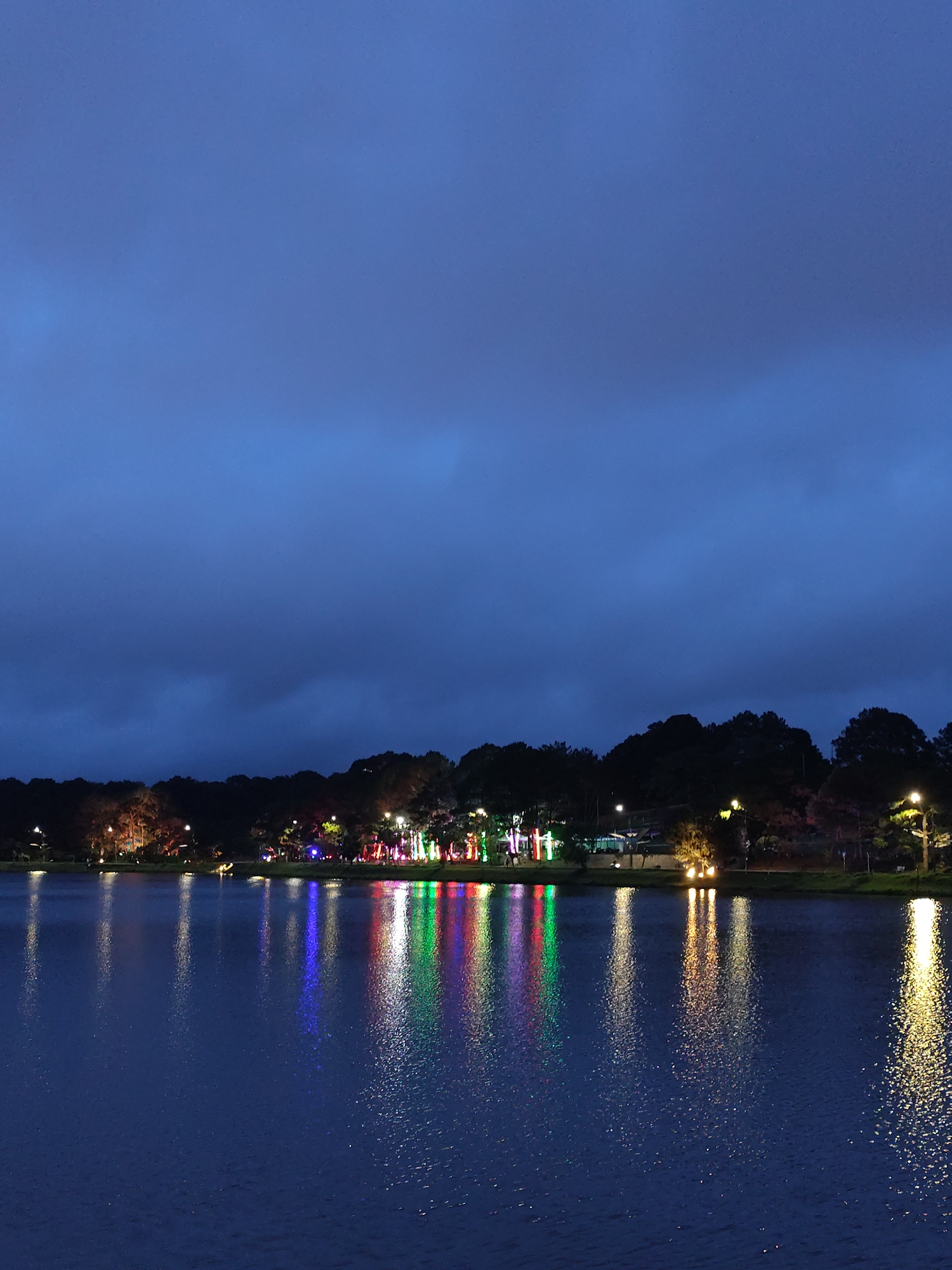 The image captures a nighttime scene at Xuan Huong Lake in Da Lat, Vietnam. The lake's dark water reflects the lights along the shoreline. Trees and buildings are visible in the background. A row of colorful decorative lights, primarily red, green, and white, stands out among the other illuminated areas, creating distinct vertical reflections in the water. The sky is a dark blue, indicative of night. No people are clearly visible.