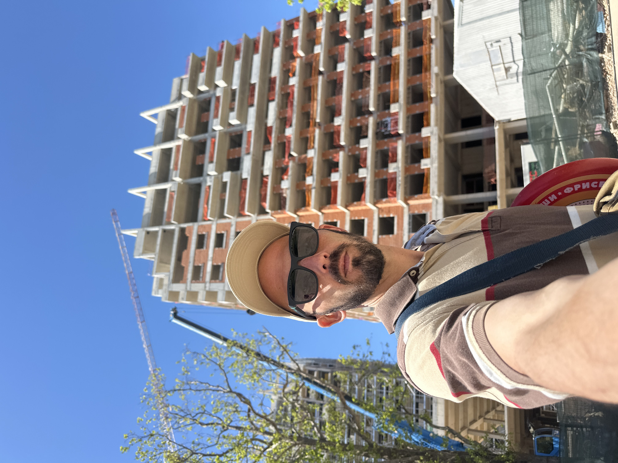 The image features a man taking a selfie outdoors, with a prominent construction site in the background under a clear blue sky. The man, positioned in the foreground, is light-skinned and bearded, wearing black sunglasses, a beige baseball cap, and a polo shirt striped in beige, brown, red, and white. A dark blue strap crosses his chest. Near his right shoulder, a red circular object, likely a frisbee, displays faint Cyrillic text that appears to read "ШИ • ФРИС".
Behind the man, a multi-story building is under construction, revealing its concrete framework. The facade features numerous rectangular openings, presumably for windows or balconies, some of which contain orange safety netting or materials. To the left, a tall construction crane rises against the sky. A smaller crane or construction vehicle is partially visible lower down, partly obscured by green tree foliage. Another structure, possibly part of the same construction project, is seen on the right, covered with mesh.
The scene takes place on a bright, sunny day, indicated by the vibrant blue sky and clear lighting. The setting is an urban development area in Budva, Montenegro. The overall impression is casual, capturing a moment against a backdrop of ongoing construction.