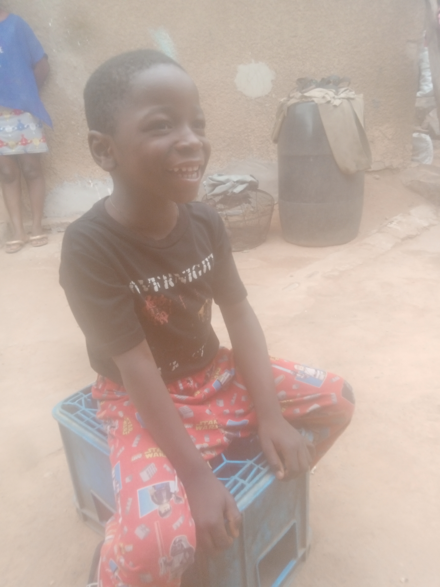 The image shows a young, dark-skinned boy sitting cross-legged on a blue plastic crate in an outdoor setting, presumably in Sabon Tasha, Nigeria. He is wearing a black t-shirt and red patterned pants with Star Wars characters. He is smiling and looking to his right. In the background, there is a blurry image of a child wearing a blue top and a patterned skirt, a large black container partially covered with a beige cloth, and a wicker basket. The ground is light brown. The image appears to be taken in daylight.