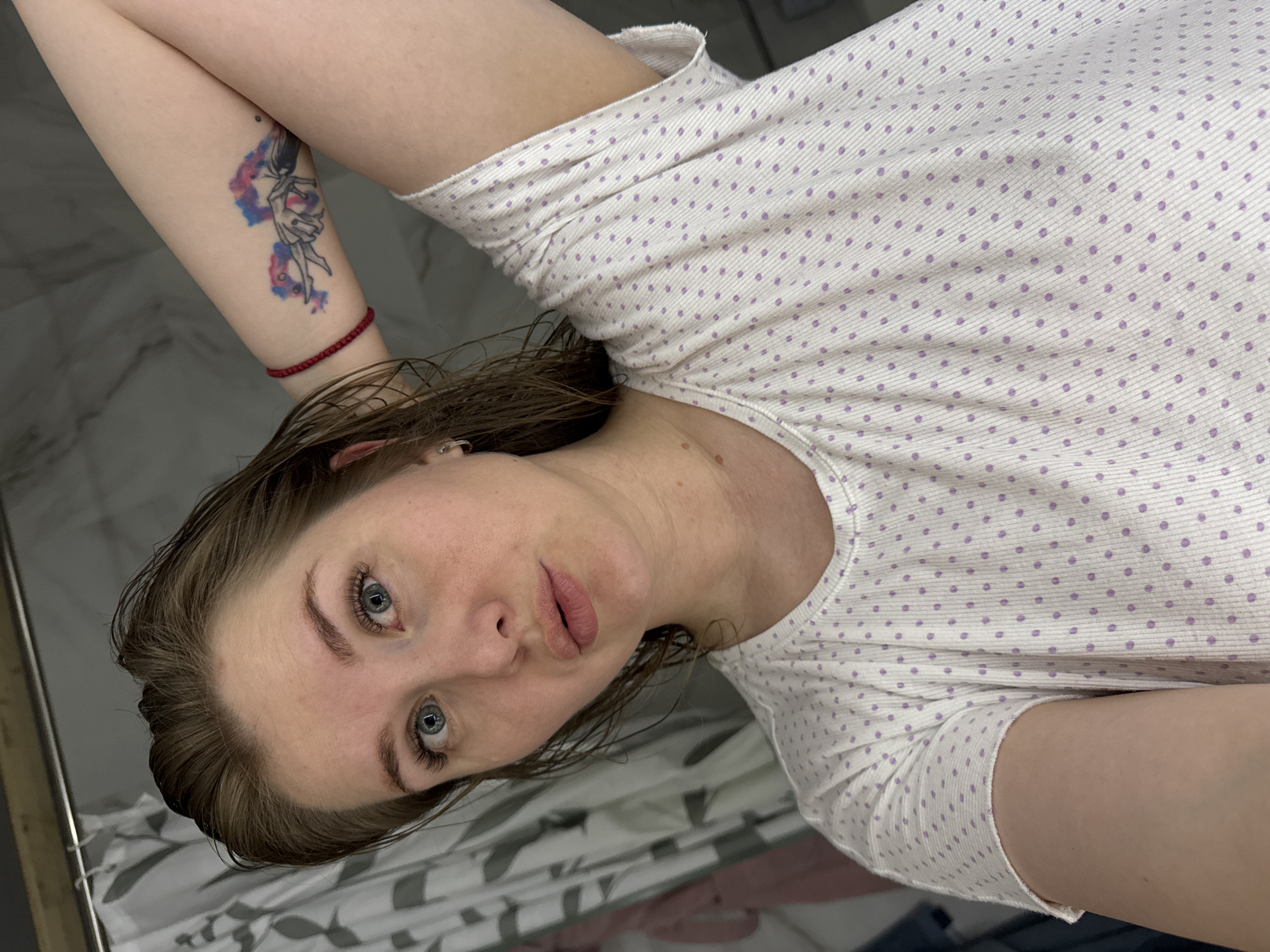 The image is a selfie taken by a fair-skinned woman with wet, light brown hair and blue eyes. She is wearing a white shirt with small purple polka dots and has a tattoo on her right inner forearm depicting a blue and red figure. A thin red string bracelet is also visible on her right wrist. She is lying down, and the background appears to be a bathroom, with a marble-like tiled floor and a shower curtain with a gray and white pattern visible in the lower portion of the frame. The location of Gornyi (Dvurechenskoe S/P), Russia is not identifiable from this image.