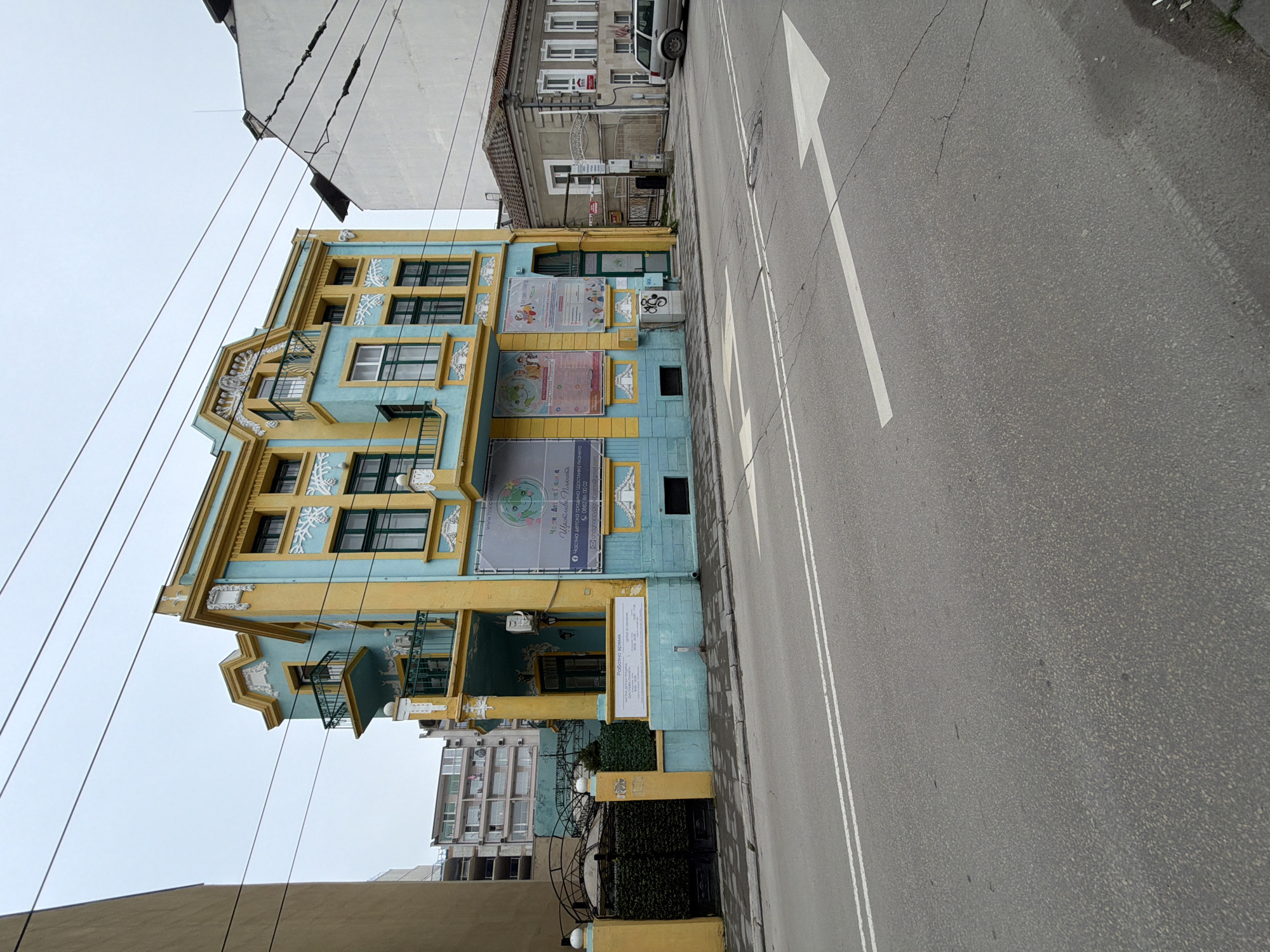 The image captures a colorful building in Varna, Bulgaria, adjacent to a road with a painted directional arrow. The building is painted in blue and yellow. It has several windows and balconies, along with signs and informational posters displayed on its facade. Power lines stretch across the upper portion of the frame. The road is grey asphalt with double white lines marking the lanes. The sky is overcast. Other buildings of varying sizes and architectural styles are seen in the background. At the ground level, a black gate with vegetation is present.