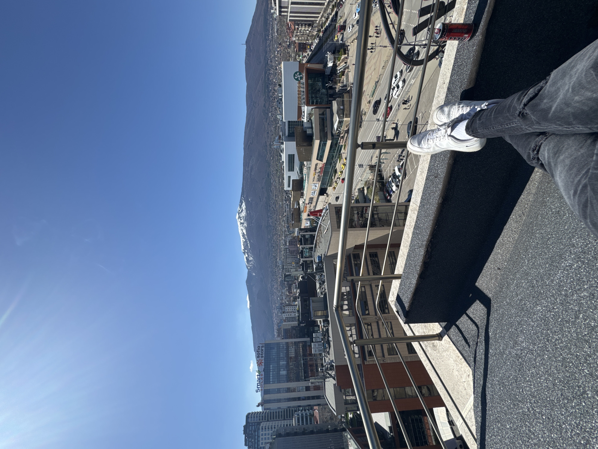 A high-angle, long shot taken in Sofia, Bulgaria from a balcony or rooftop shows the city skyline, a mountain range, and a person sitting with their legs crossed. The person is wearing white sneakers and grey jeans with rips at the knees. A can of Coca-Cola sits on the edge of the balcony. The city includes visible buildings, streets with cars, and the "Paradise Center" shopping mall which has a Starbucks logo. The mountains are in the background and have patches of snow on them. The sky is blue and clear. The photo is taken during the daytime, as suggested by the bright light and shadows.