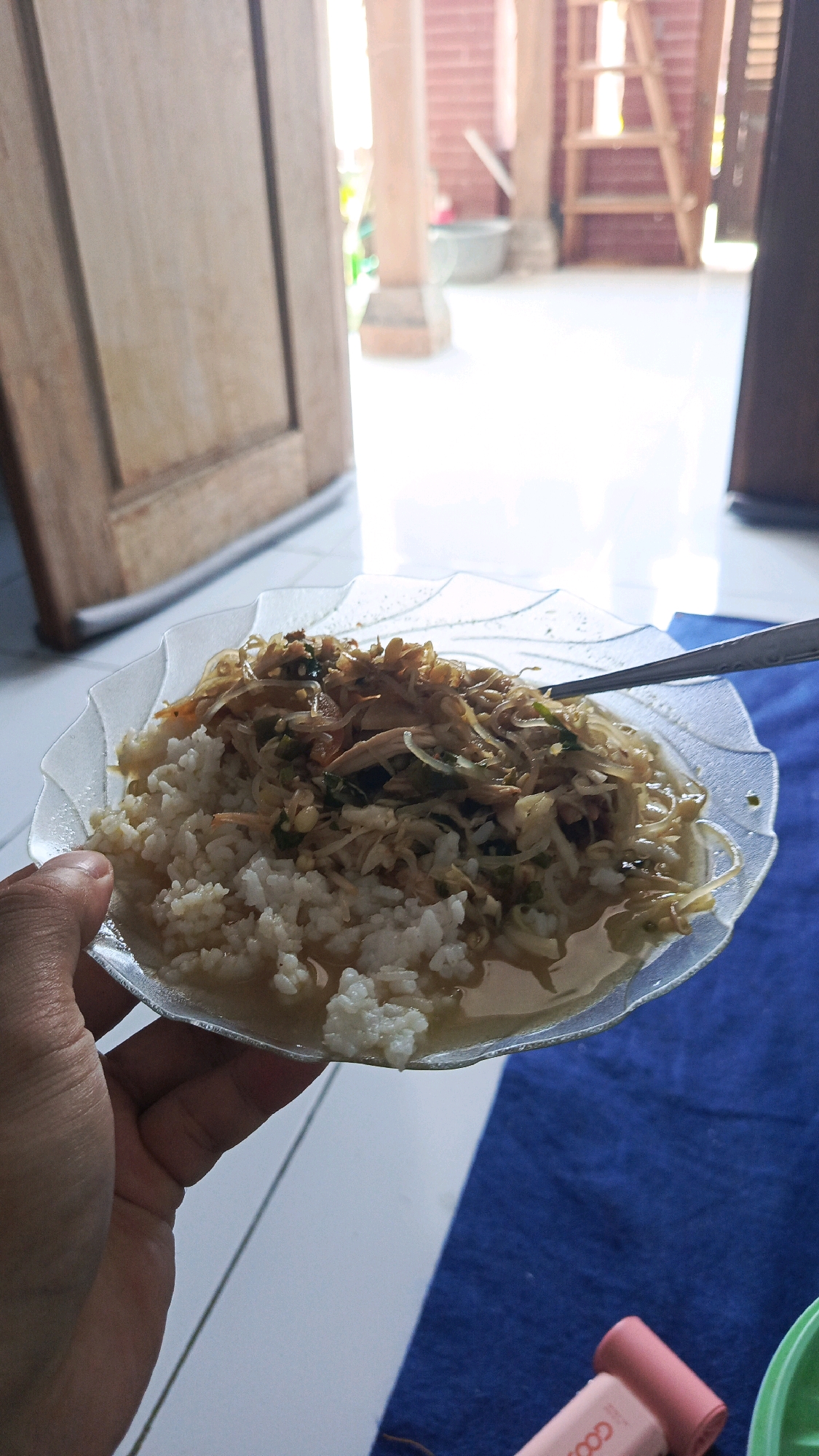 A person's hand is holding a clear, scalloped-edge plate of food. The plate contains white rice at the bottom, topped with a mixture of shredded chicken, bean sprouts, and herbs in a light broth. A metal spoon rests in the food. The plate is positioned over a textured blue surface, likely a fabric. In the background, a wooden door is visible on the left, and a brick wall with vertical wooden beams and a wooden ladder are present on the right. The setting appears to be an outdoor or semi-outdoor area in Babadan, Indonesia, possibly a porch or patio, with bright natural light.